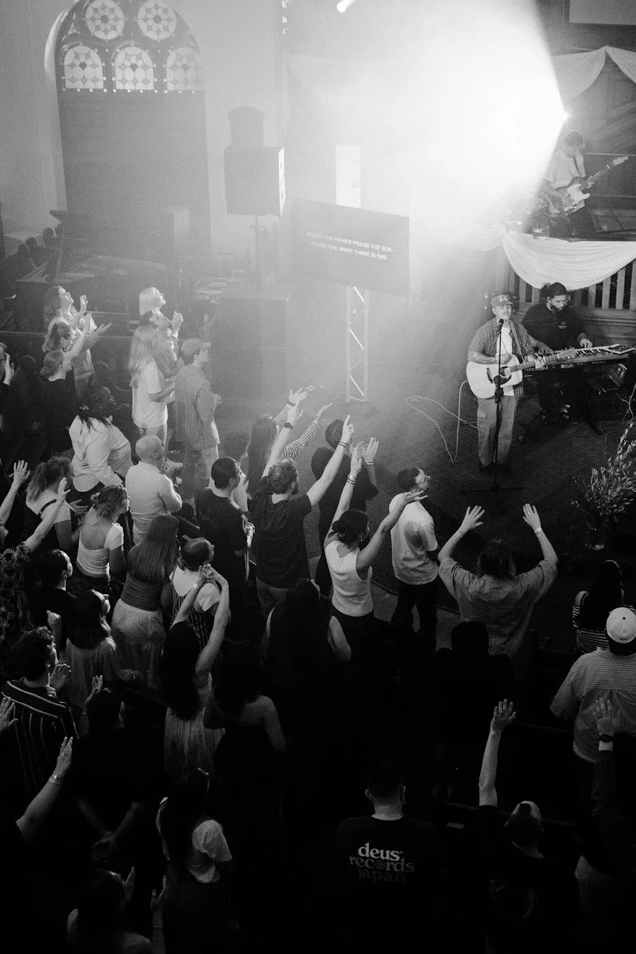 A black and white photo of a church concert with a crowd standing and raising their hands, a guitarist singing into a microphone, and a keyboard player nearby. The church interior features stained glass windows.