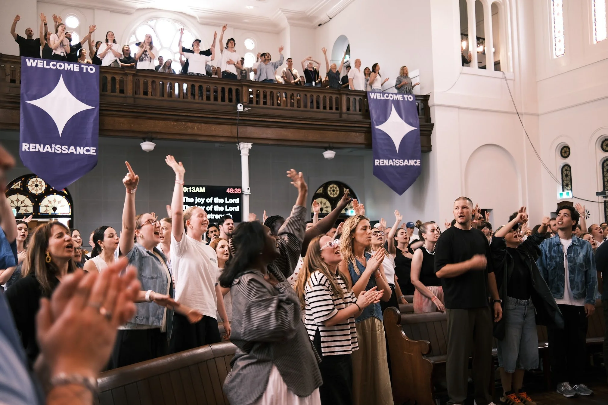 A large congregation of people inside a church, participating in a religious service. Some are standing, raising hands, and singing. Two purple banners with the words 'Welcome to Renaissance' hang from the balcony, which has more people watching from above. The church has high ceilings, stained glass windows, and wooden pews.