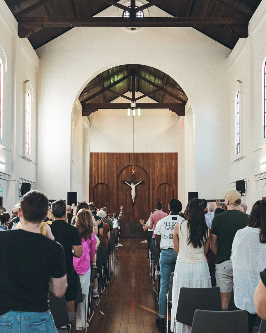 People inside a church attending a religious service, with a crucifix on the wooden wall at the front.