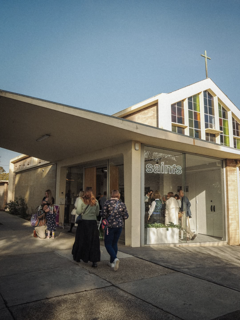 Group of people entering a church with a glass storefront marked 'saints' in a suburban area, modern church building with stained glass windows and a cross on top.