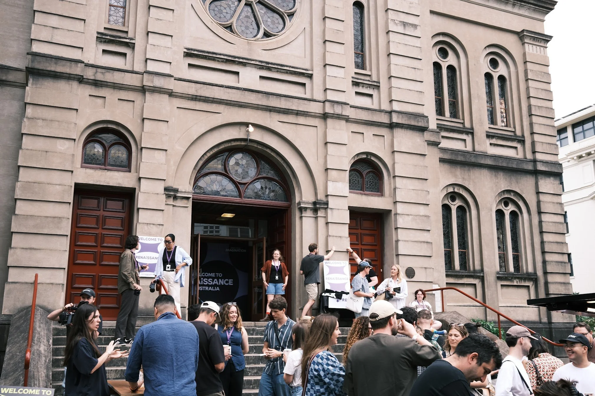People gathering on the steps of a historic church or cathedral with large wooden doors and stained glass windows, during daytime.
