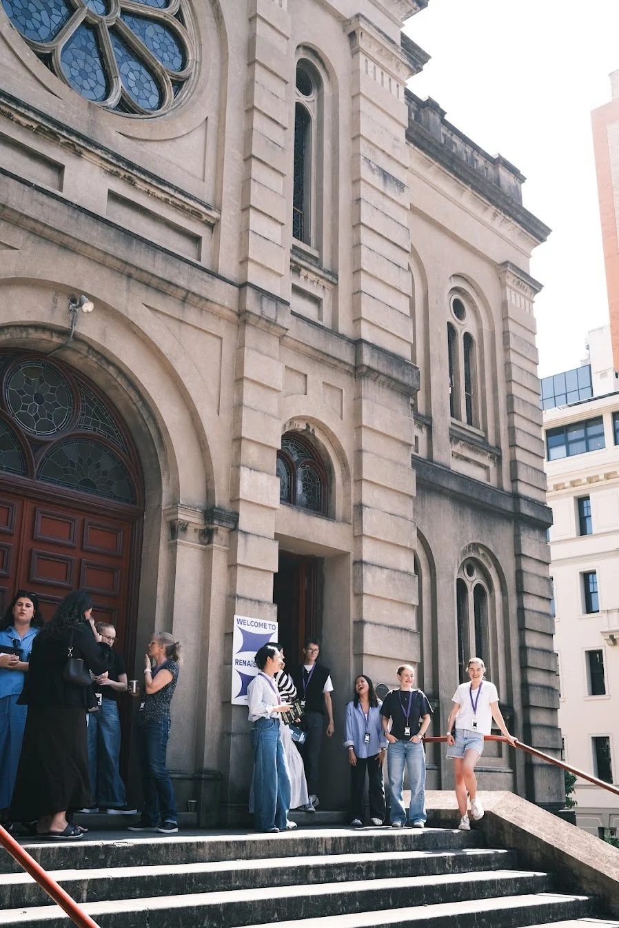 Group of people standing and sitting on the steps outside a historic stone building with large arched windows, some wearing lanyards. A sign that reads 'Welcome to Renaissance' is visible near the entrance.