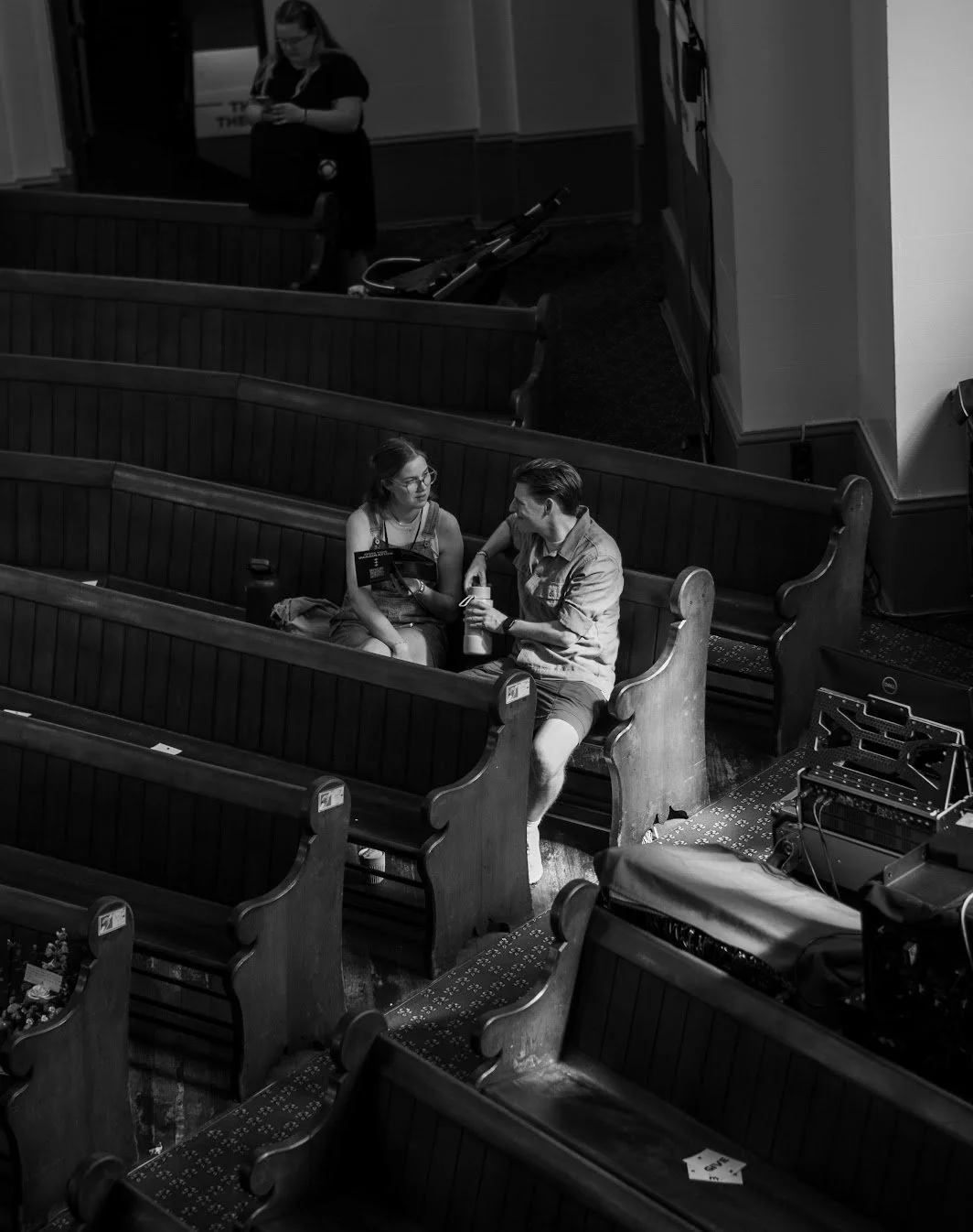 Two people sitting in a church pew, engaged in conversation, with one holding a drink. A woman stands in the background looking at her phone, and there are empty pews around them.