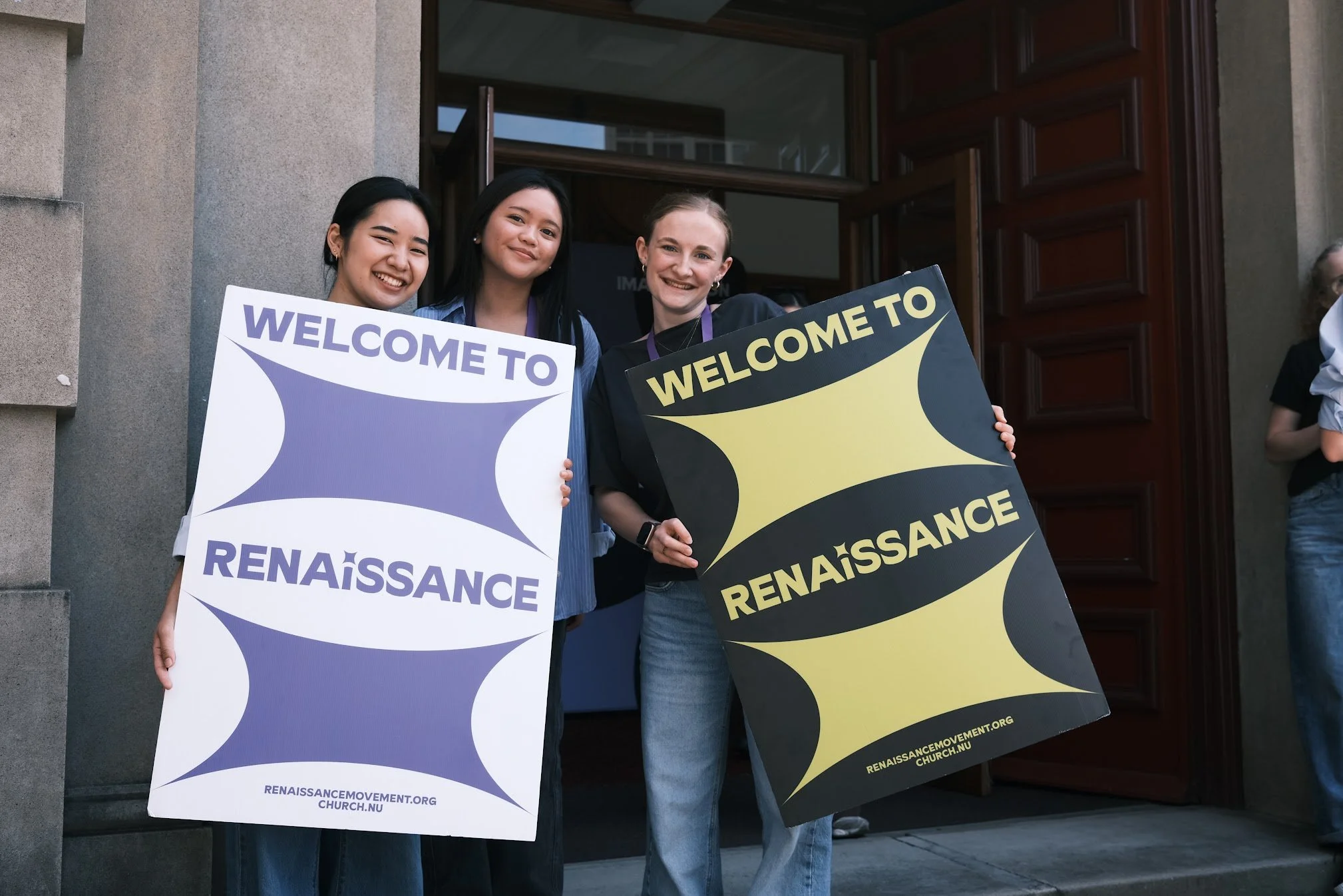 Three young women standing outside a building, holding signs that say "Welcome to Renaissance" with the website URL at the bottom, smiling and posing for the photo.