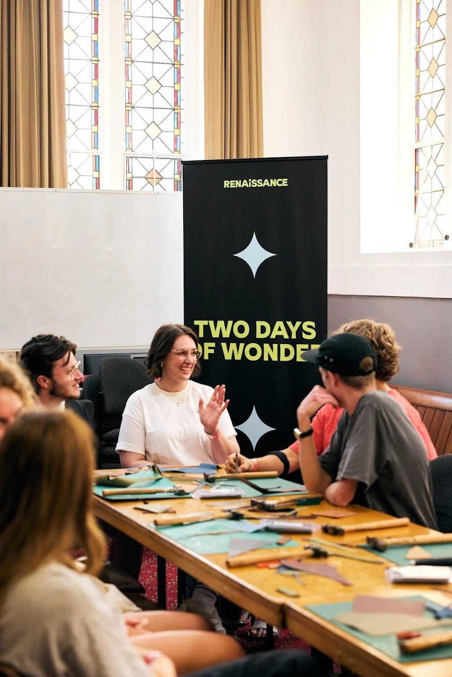 A group of people sitting around a wooden table, engaged in conversation and smiling. Behind them is a black banner with yellow text that reads "Renaissance Two Days of Wonder." The room has large stained glass windows and beige curtains.
