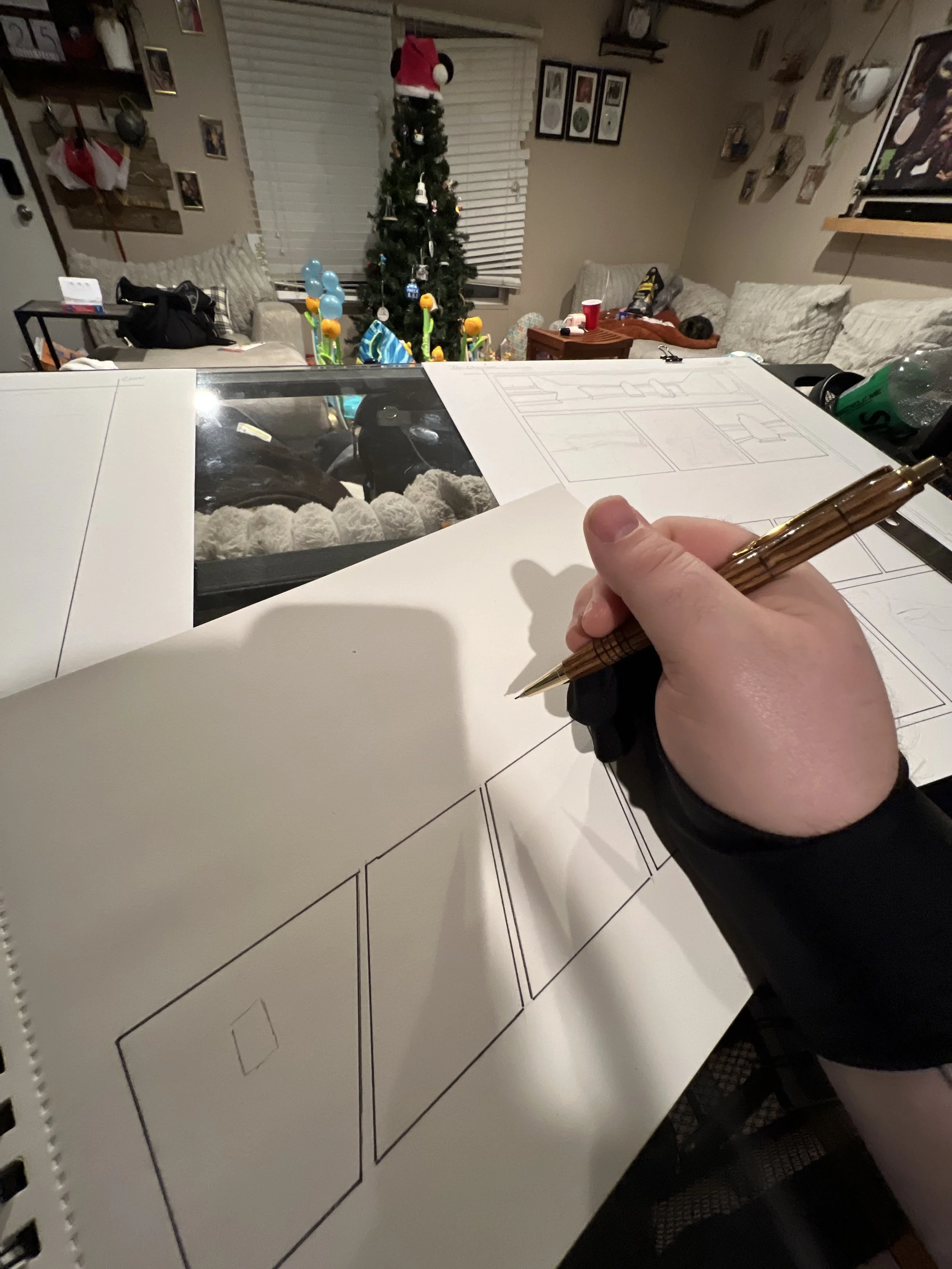 A person is drawing house floor plans on a large sheet of paper. In the background, there is a decorated Christmas tree, a couch with cushions, a small table with a red cup, and a wall with framed pictures.