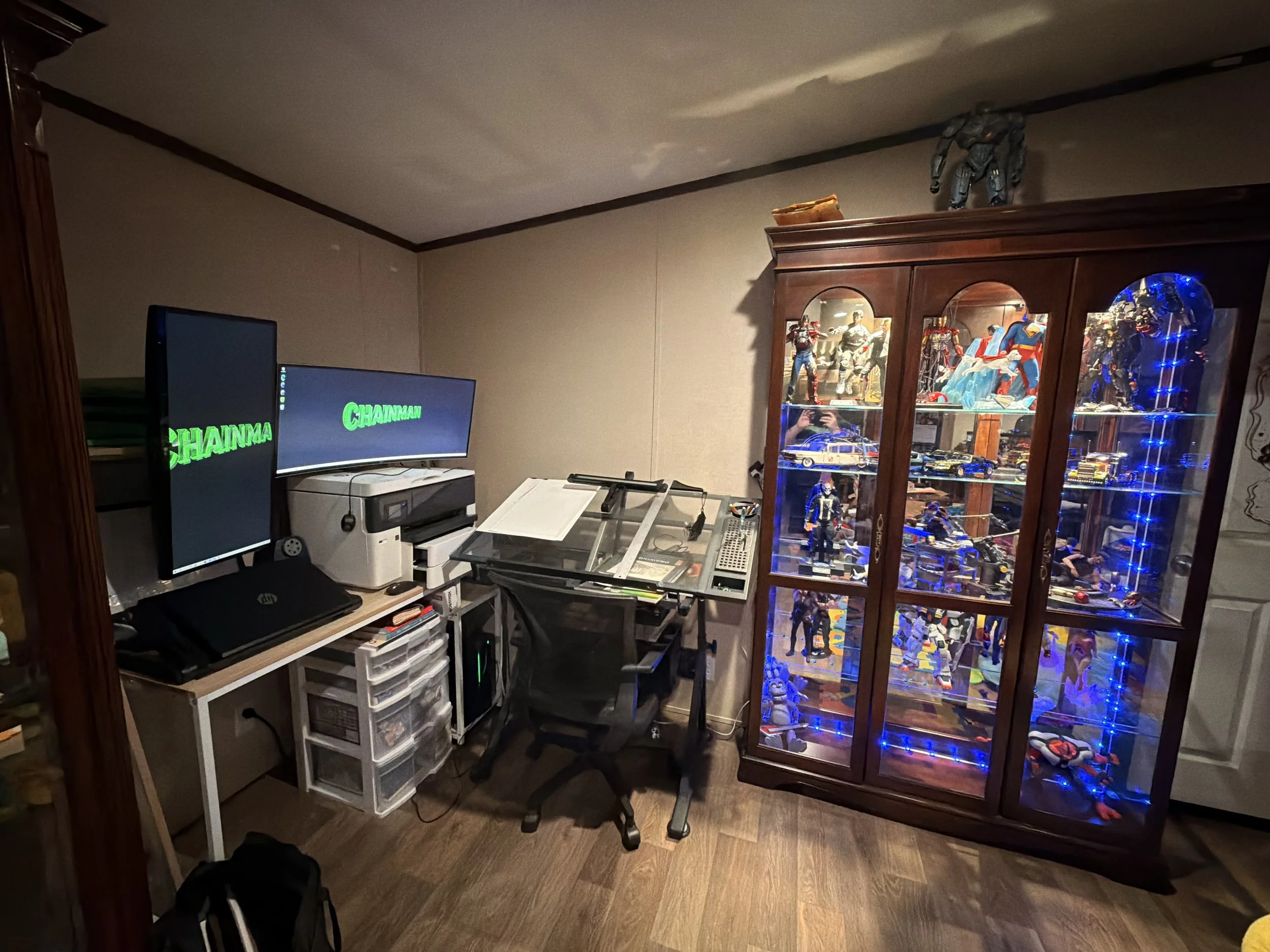 A room containing two computer monitors, a white printer, a desk with papers and a gun on it, a black swivel chair, and a large wooden display cabinet filled with action figures and collectibles, with blue LED lights highlighting the items inside.