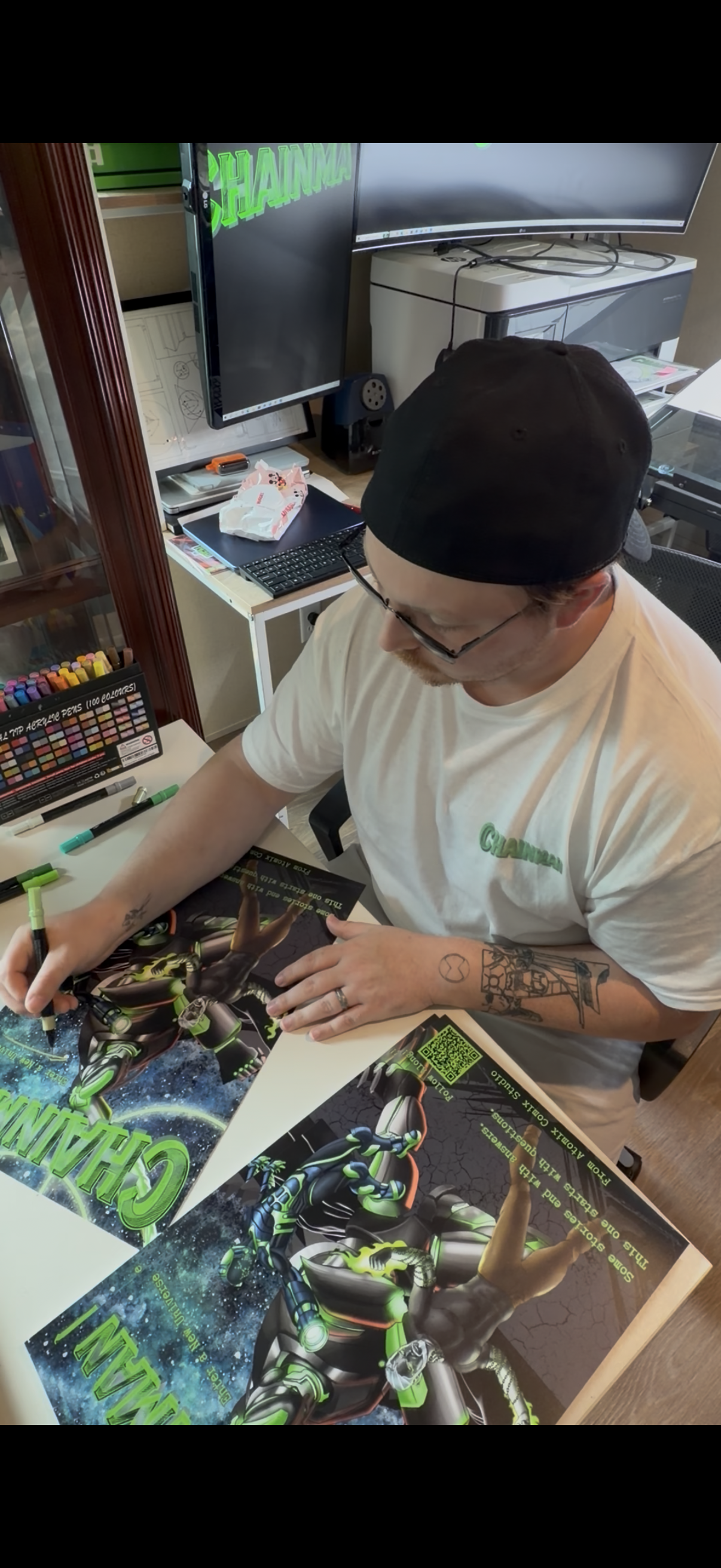 A man wearing glasses, a black cap, and a white t-shirt signing posters for a Call of Duty gaming event. The table has posters and markers, and there are two monitors in the background.