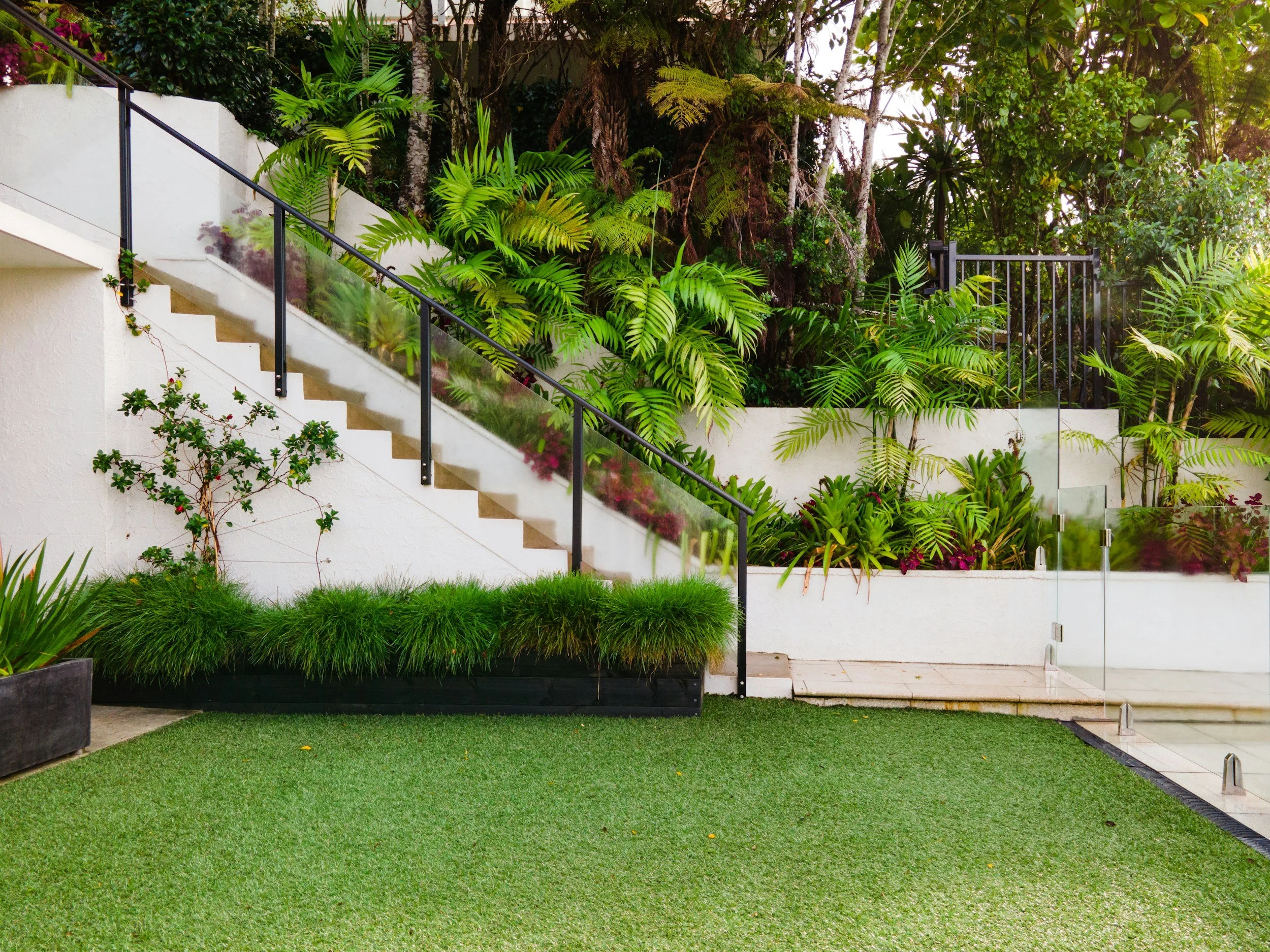 A modern outdoor area with a white staircase, a glass railing, lush green plants, and a tall hedge behind.