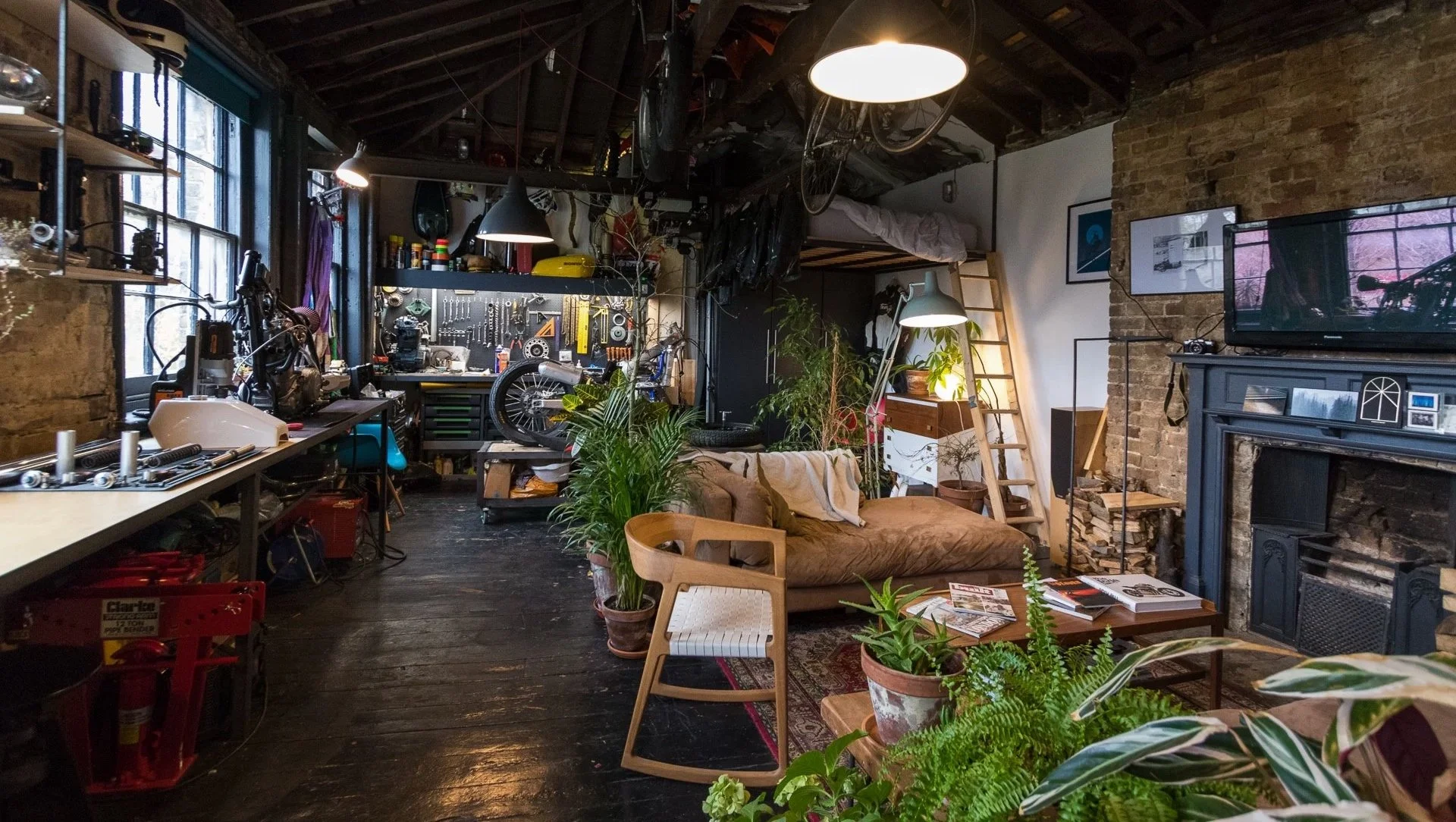 A cozy urban loft motorcycle workshop. Exposed brick walls, a brick fireplace, and a large window with natural light. multiple work areas were created. built in workbenches, architectural lighting, welding table, custom motorcycles.