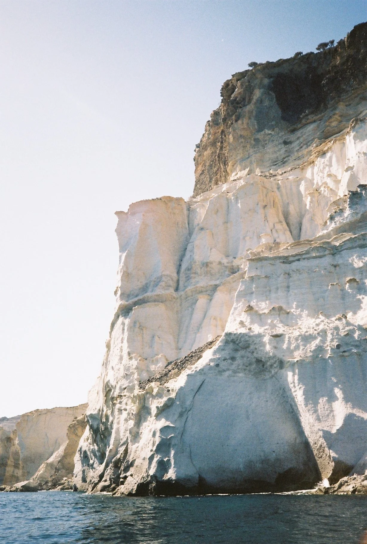 Cliffs along a coastline with white and dark rock formations and blue ocean water at the base, under a clear sky.