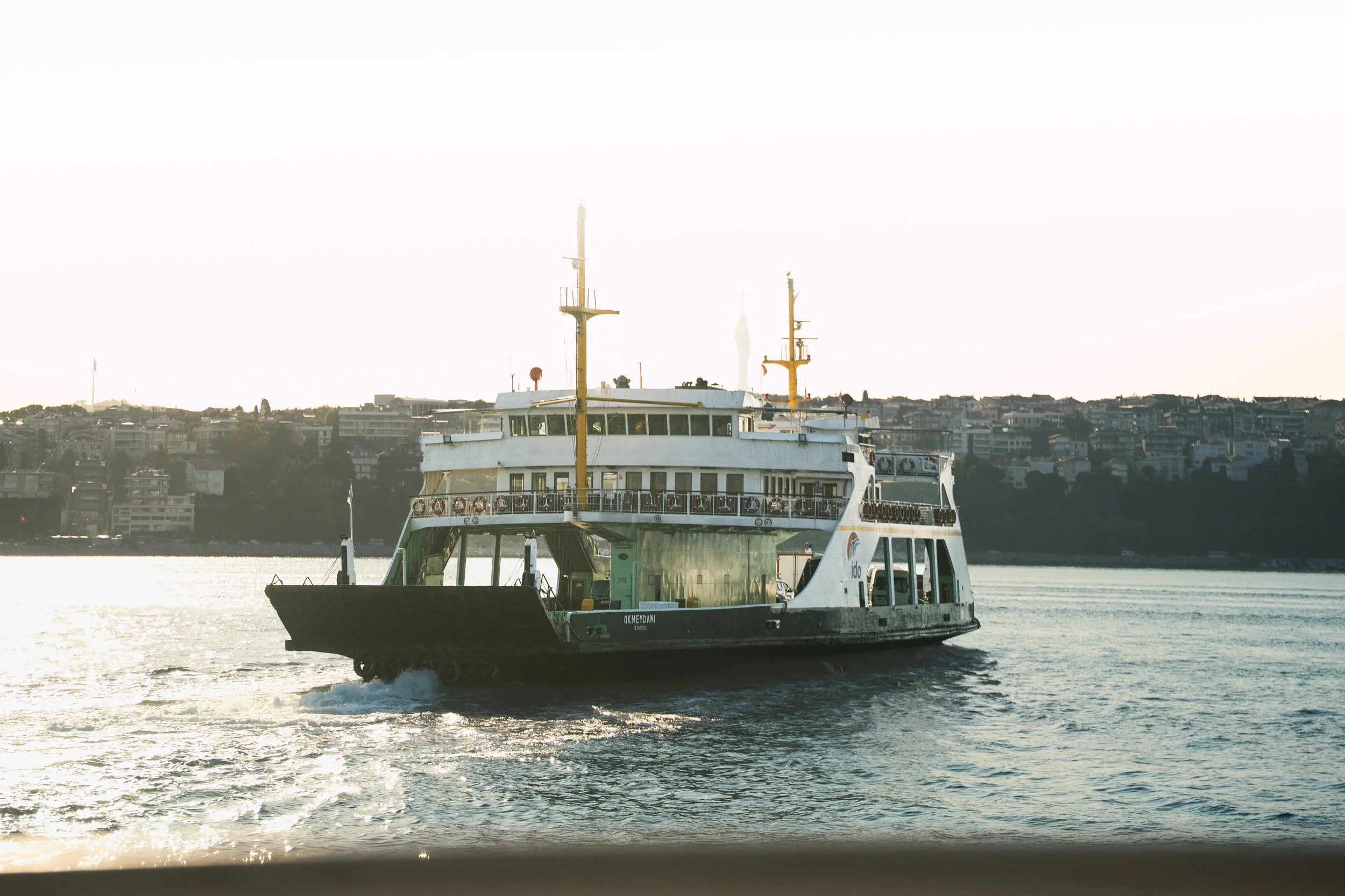 A ferry boat named OK MEYDANI sailing on water with a cityscape in the background and a clear sky.
