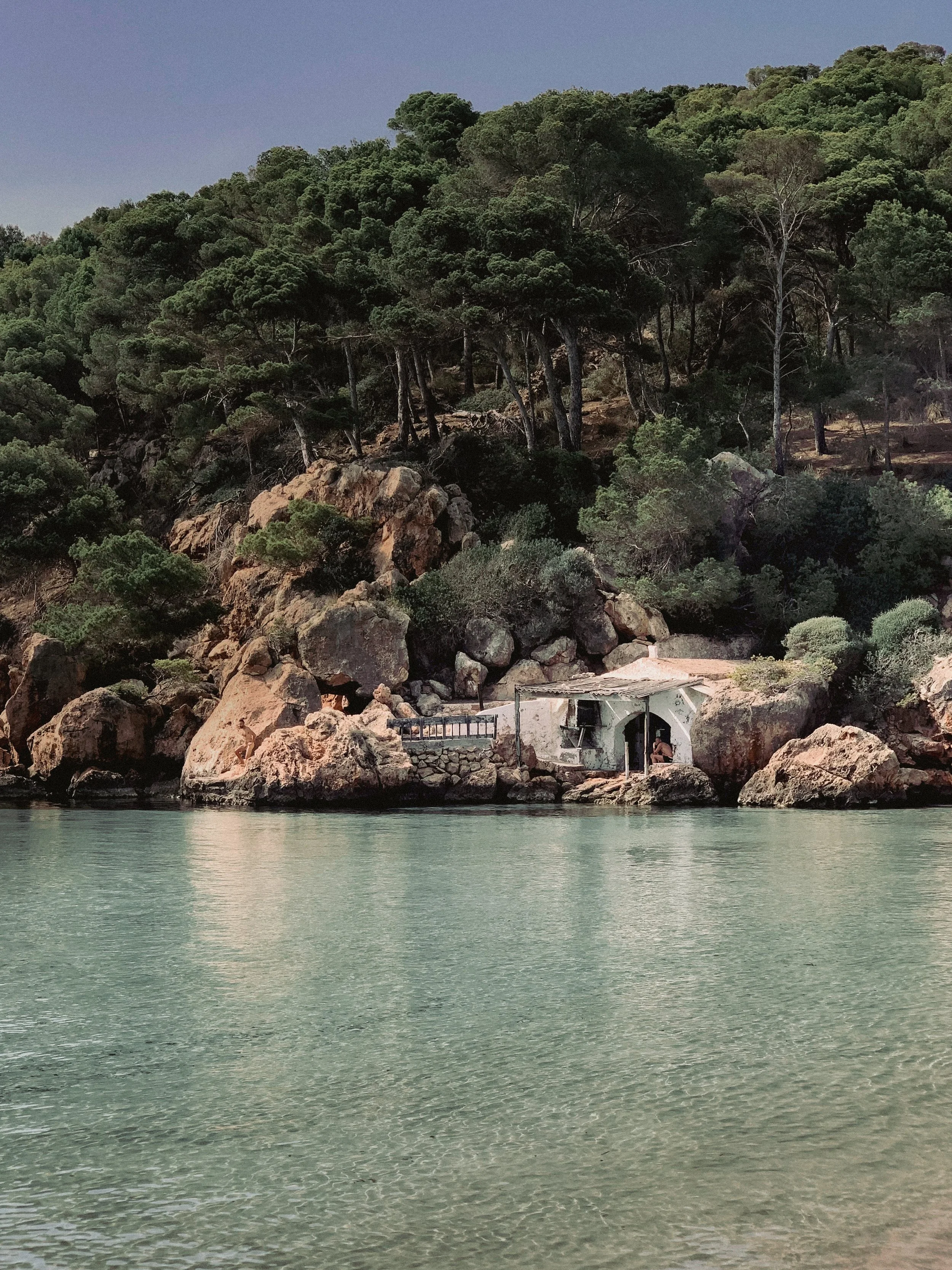A small white house with a chimney and wooden roof, built into a rocky cove along the water, surrounded by large rocks and green trees on a hillside.