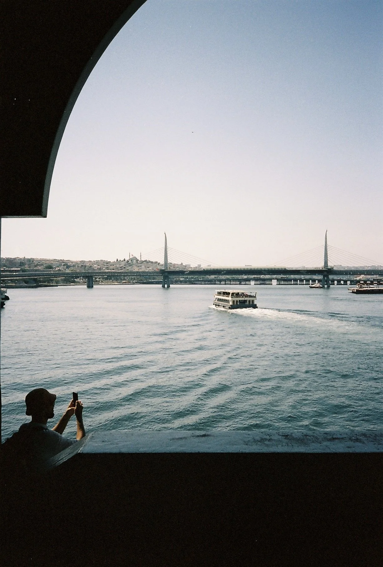 View of a river with a passenger ferry boat, bridge, and cityscape in the background, taken from inside a boat or ferry.