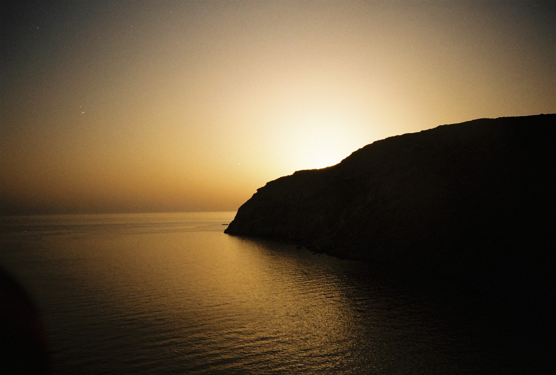 Silhouette of a large hill or mountain by the water at sunset or sunrise, with calm water reflecting the sky and a soft, warm glow on the horizon.