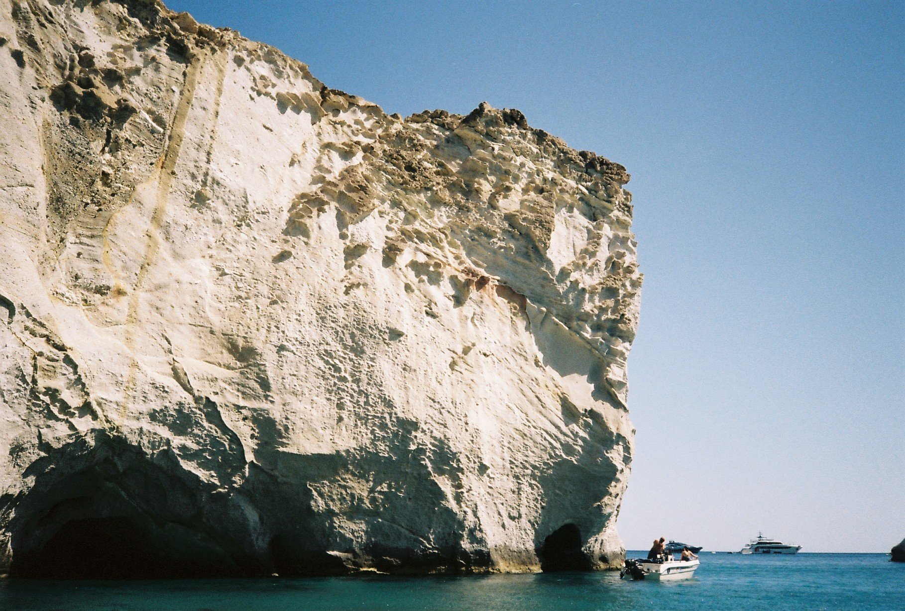 Large white and beige cliff with a small cave at the base, blue sky, boats and people in the water near the cliff.