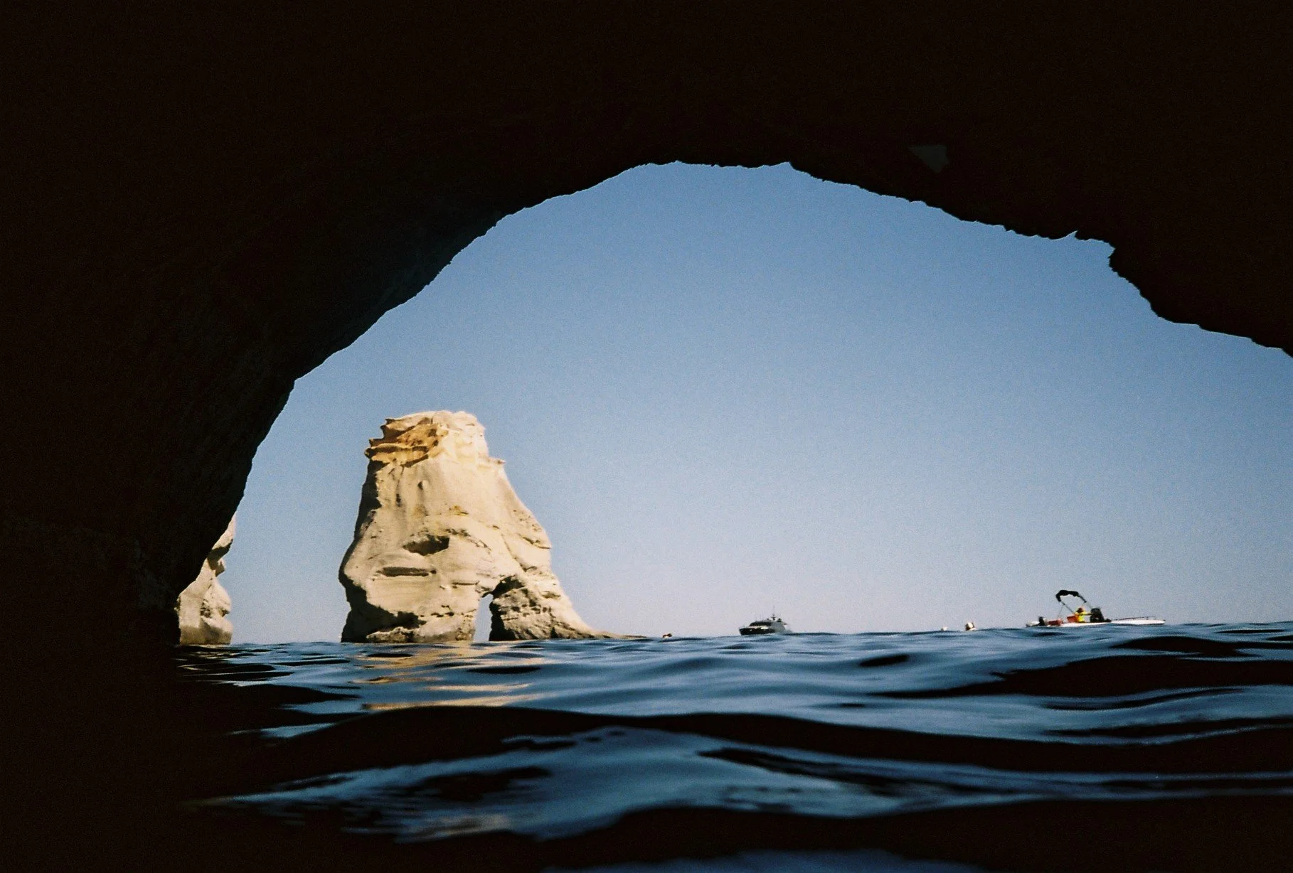 View of the sea with large rock formations, seen from underneath a natural rock arch, with boats in the distance and a clear blue sky.