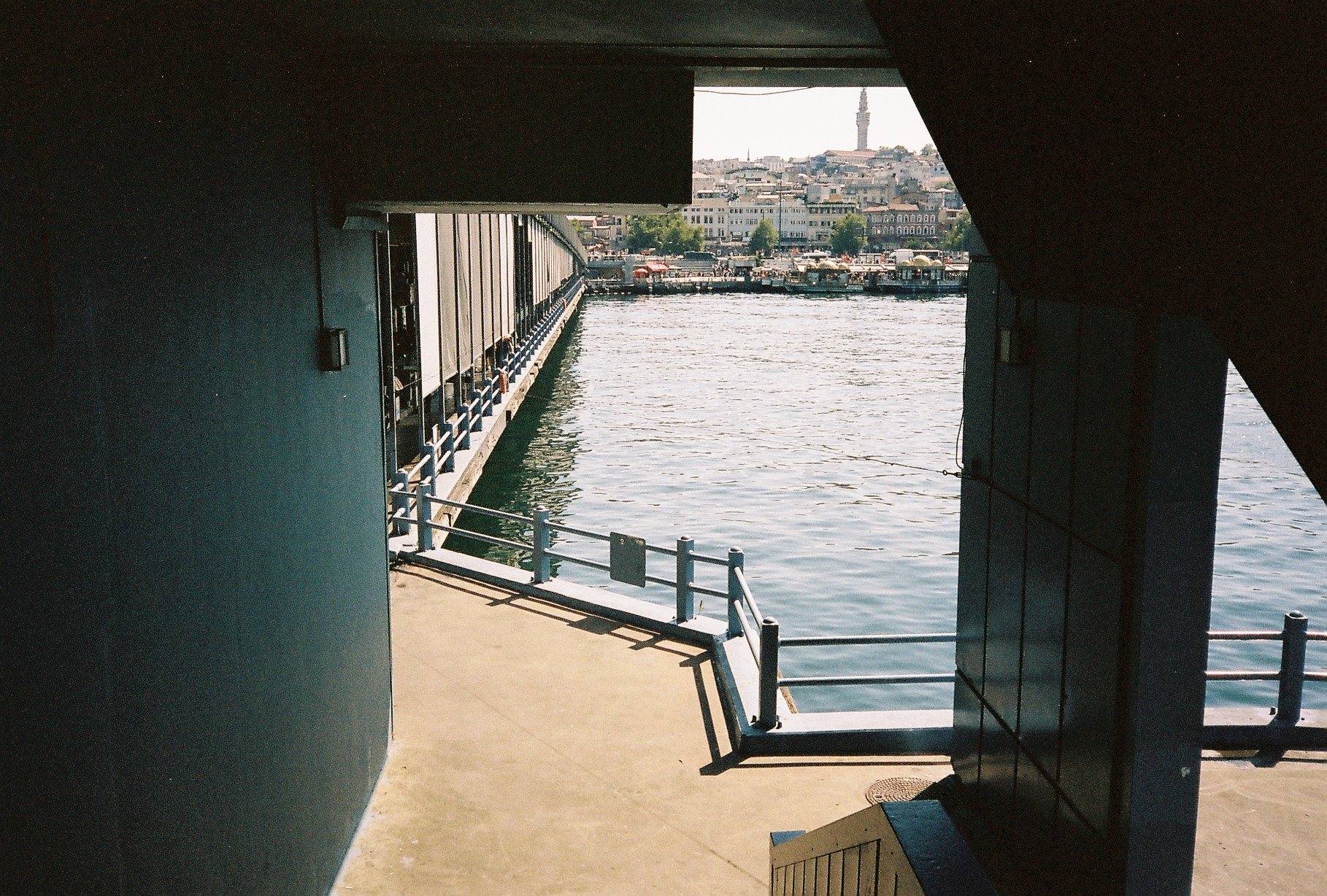 View of a waterfront scene through a dark, shadowed opening, showing water, boats, a cityscape with buildings, and a tower in the distance.