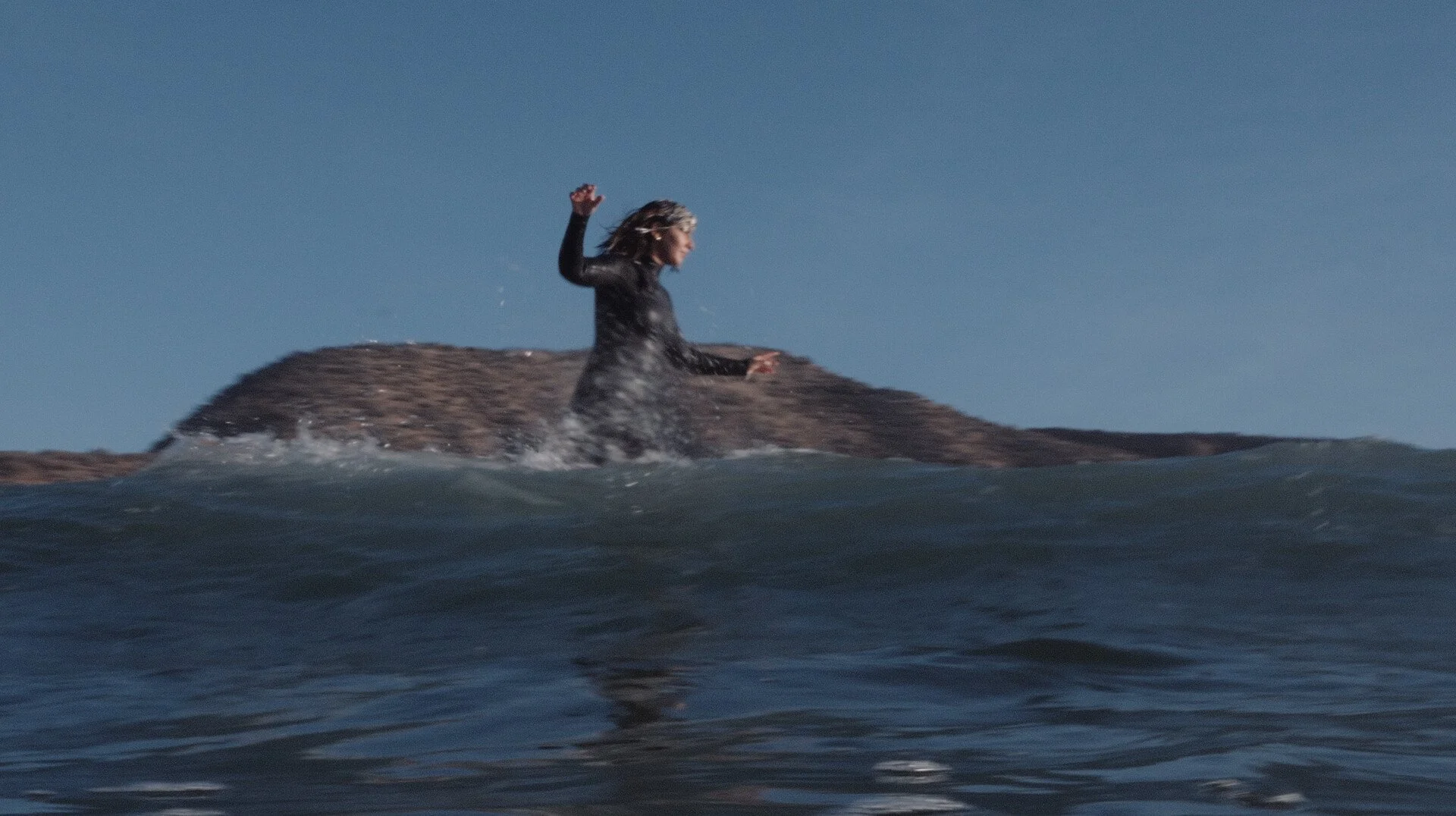 A woman in a black wetsuit sitting on a large rock floating in the ocean, waves crashing around her, under a clear blue sky.