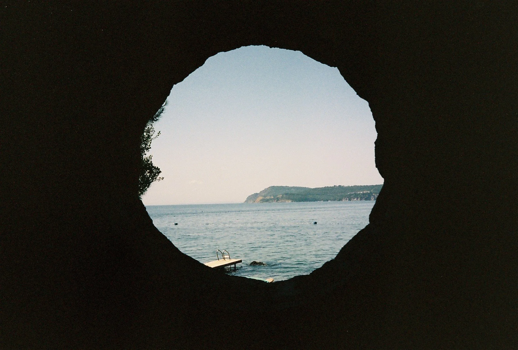 View of the sea and distant coastline seen through a cave opening, with a swimming pool ladder and dock in the foreground.