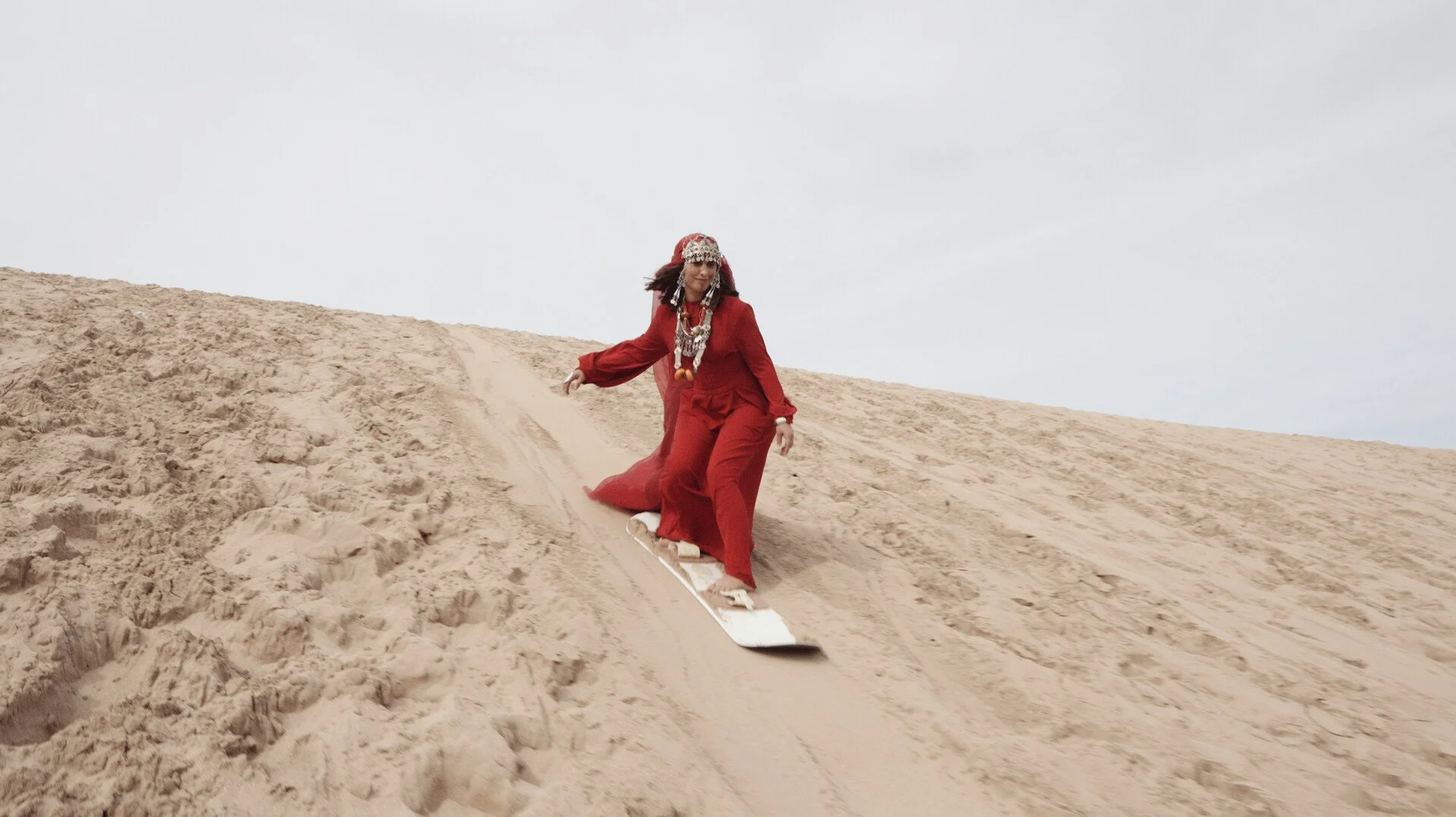Woman in a red dress and jewelry skiing down a sandy hill.