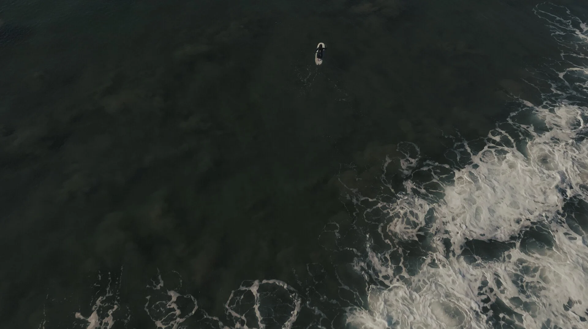 An aerial view of a person on a surfboard in the ocean, with waves and foam visible around them.