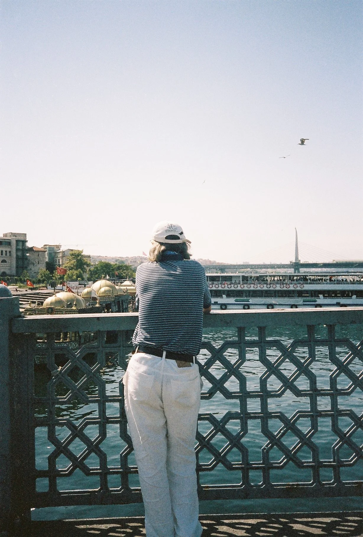 Person with white pants, striped shirt, white cap, and sunglasses leaning on a bridge railing, overlooking a river with boats, buildings, and a modern suspension bridge in the background, and seagulls flying in the sky.