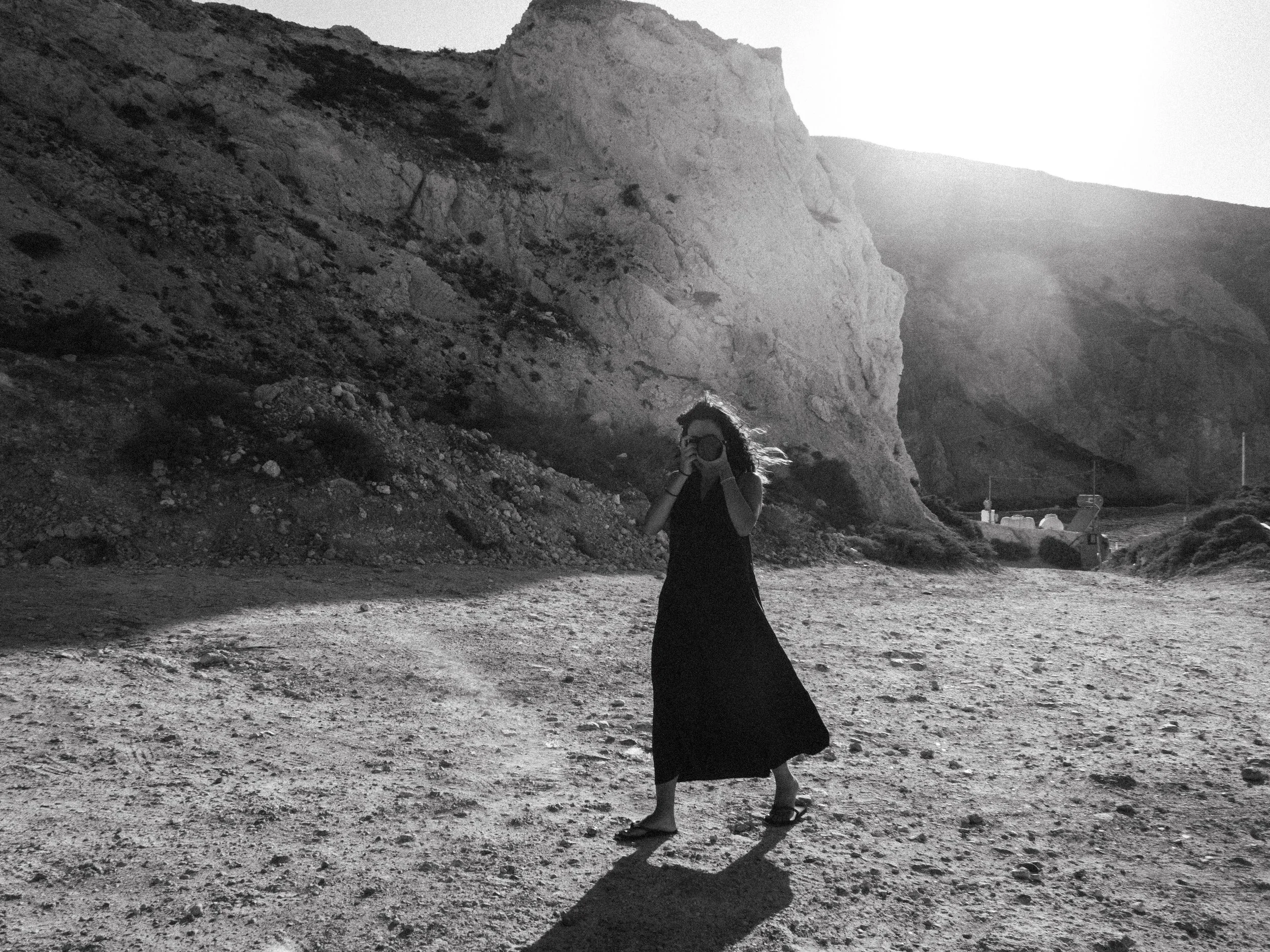 A woman with long hair wearing a black dress and sandals standing on rocky terrain near cliffs, holding sunglasses, with mountains and bright sunlight in the background.