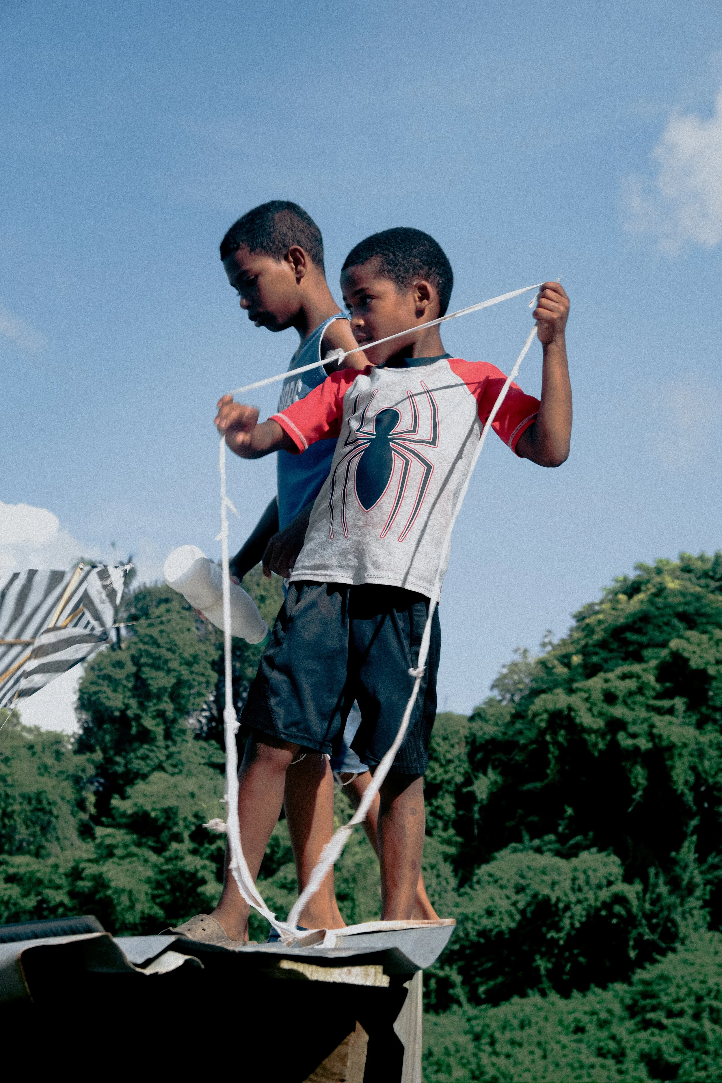 Two children standing on a platform outside against a blue sky and green trees, one wearing a gray shirt and the other a red and gray shirt with a spider emblem, holding a string connected to a model boat.