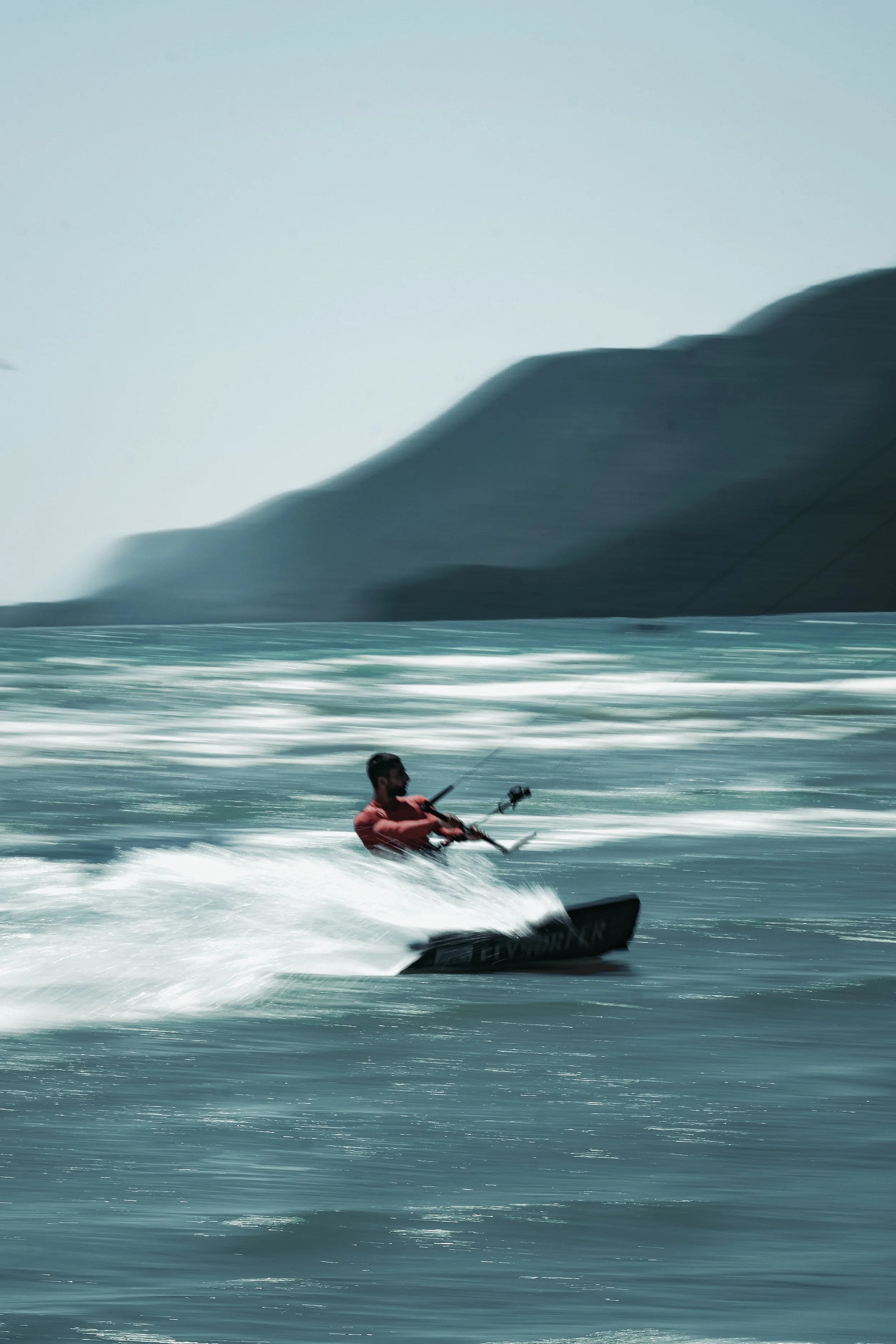 A person in a red wetsuit riding a jet ski at high speed on the ocean, with a large cliff or mountain in the background.