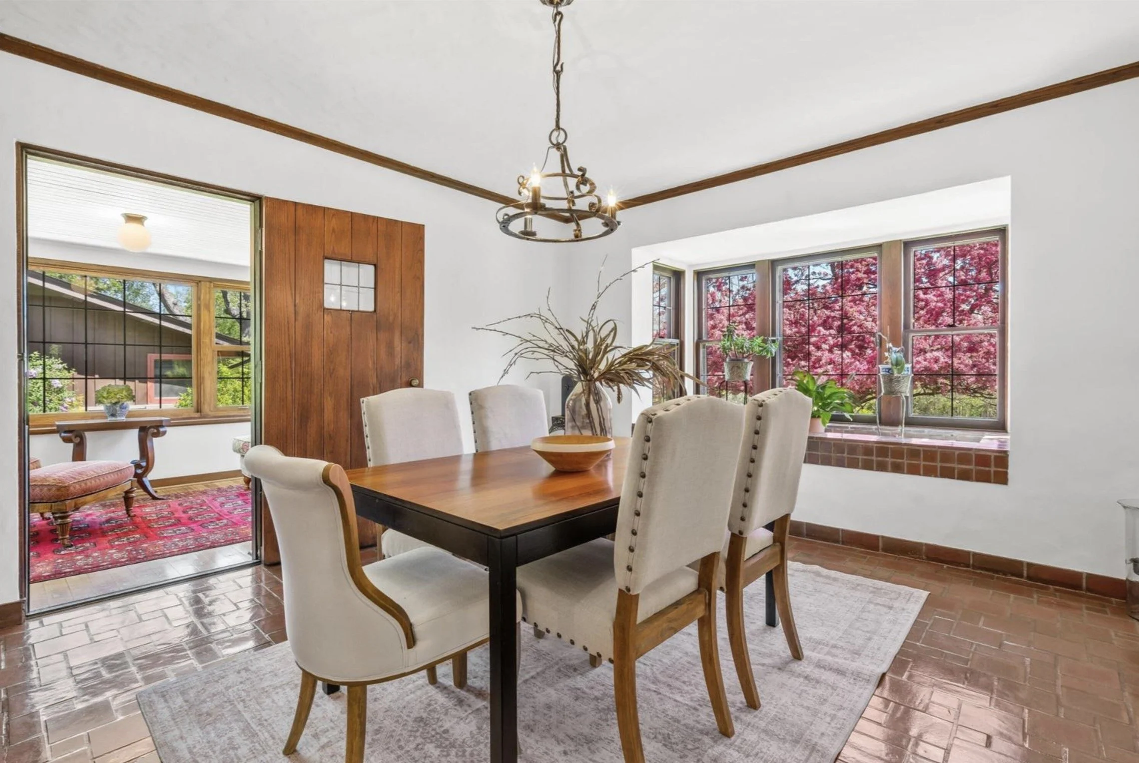 A dining room with a wooden table, cushioned chairs, and a decorative centerpiece. Large windows show trees with pink foliage outside. The room has a white ceiling with wooden trim and a rustic chandelier.