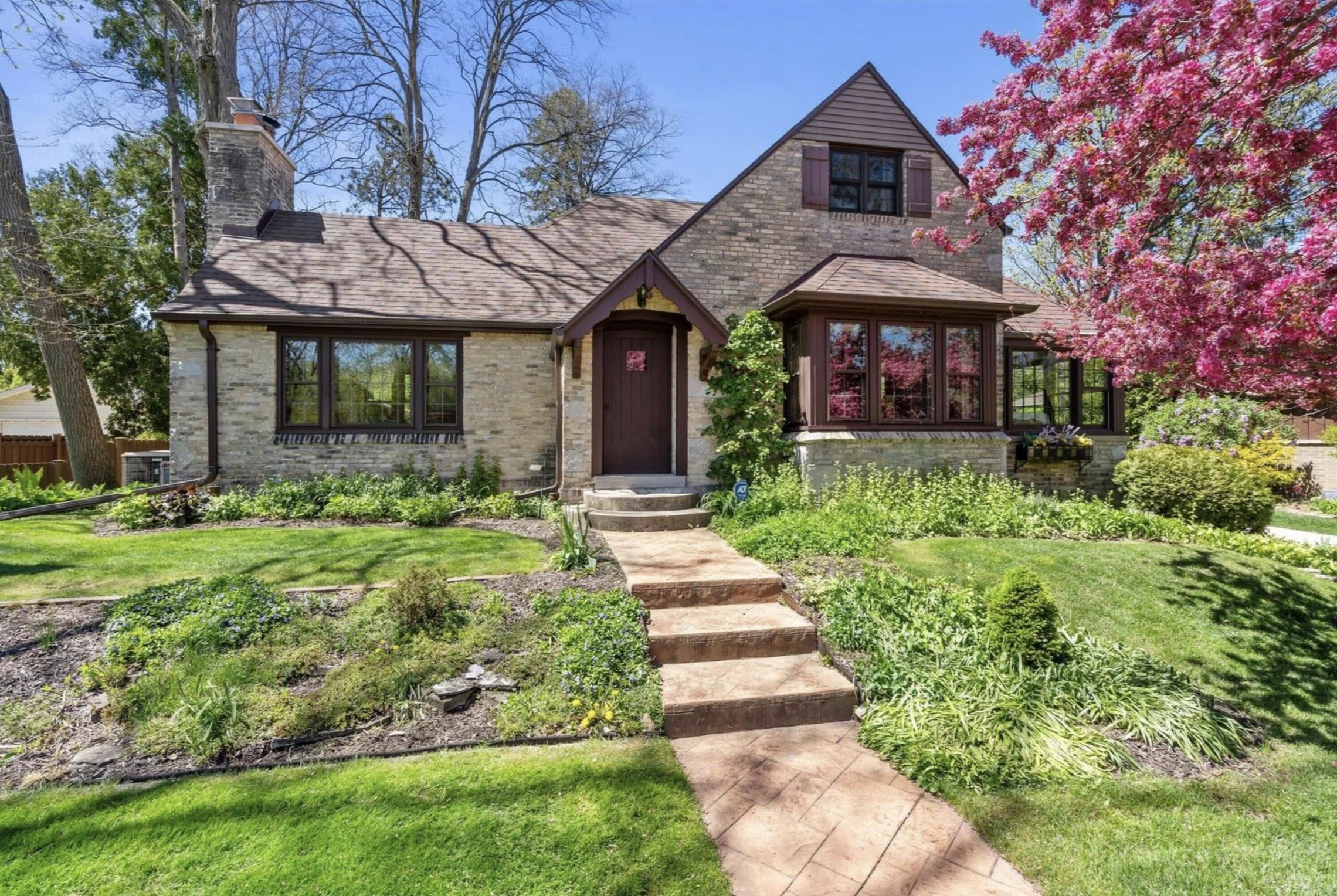 Brick house with a brown front door, surrounded by a lush green lawn, trees, and pink flowering tree on the right side.