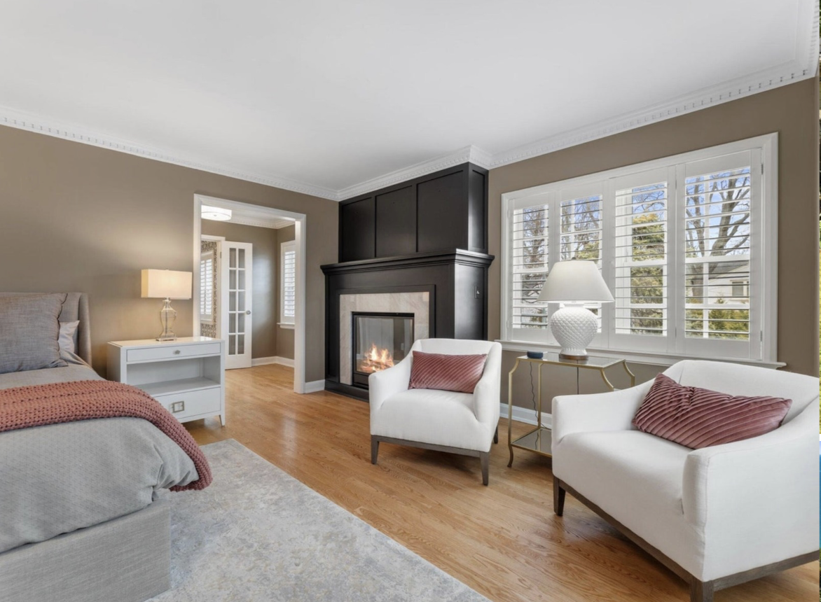 Living room with a fireplace, two white chairs with pink pillows, a white side table with a lamp, a large window with shutters, hardwood floors, and beige walls.