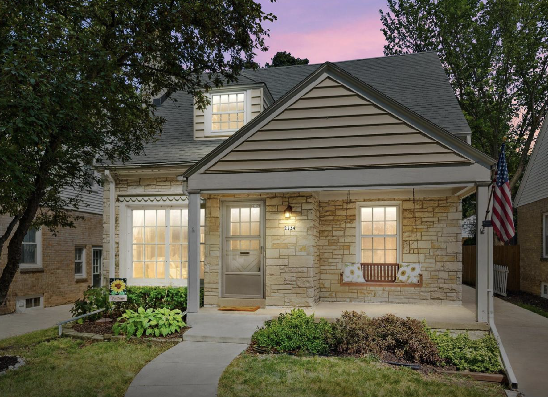 Front view of a cozy house with a stone facade, a small porch, and a front yard with a curved walkway, lush greenery, and a bench. The house has a dormer window on the roof, warmly lit inside, with an American flag hanging on the porch. The sky in the background is pink and purple during sunset.