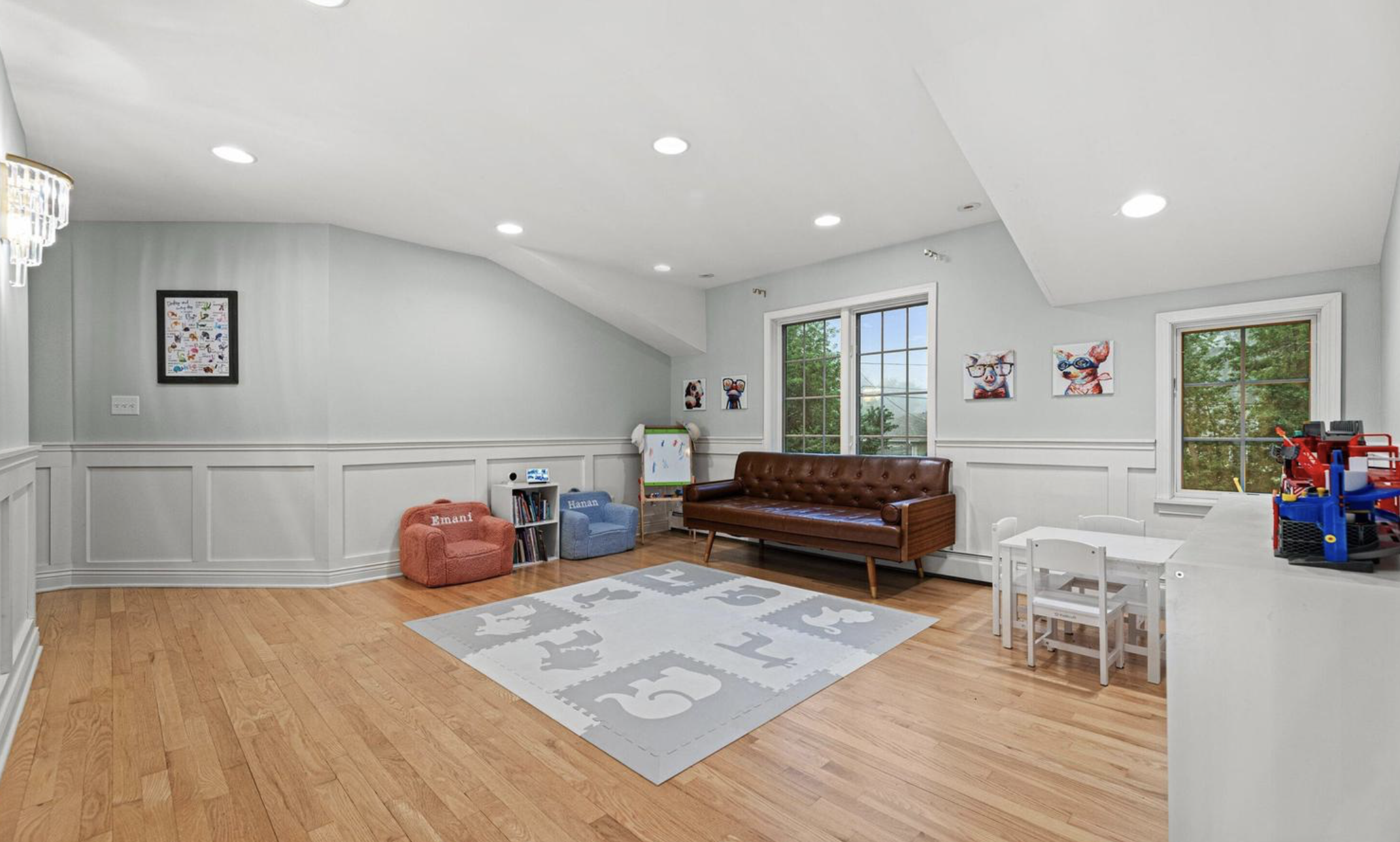 A cozy children's playroom with hardwood floors and light gray walls. There are two small upholstered armchairs, one pink labeled "Emani" and one blue labeled "Hanan." A white bookshelf with children's books and a small whiteboard are nearby. On the wall are two colorful animal art prints. Two large windows let in natural light. There is a brown leather sofa, a white table with chairs, and various children's toys, including a red and blue plastic playset.