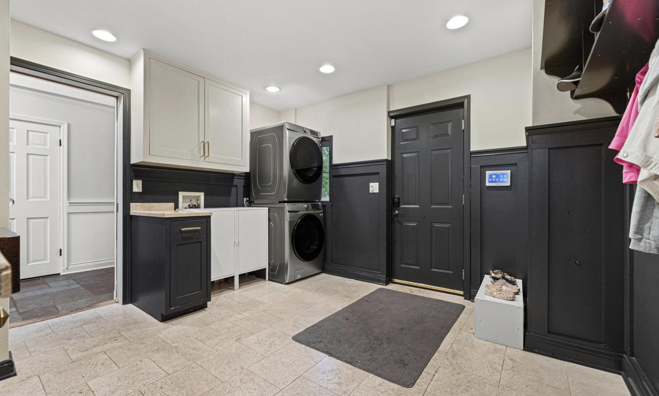 Laundry room with stacked washer and dryer, white and black cabinets, black door, and tiled floor.