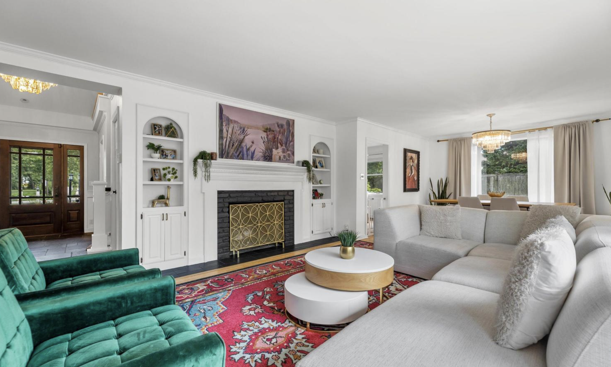 Living room with white walls, a white sofa, green velvet armchair, and a red patterned rug. Fireplace with a black brick surround, gold decorative screen, and framed art above. Built-in white shelves with books and decor. Large window with beige curtains, dining table, and chandelier.