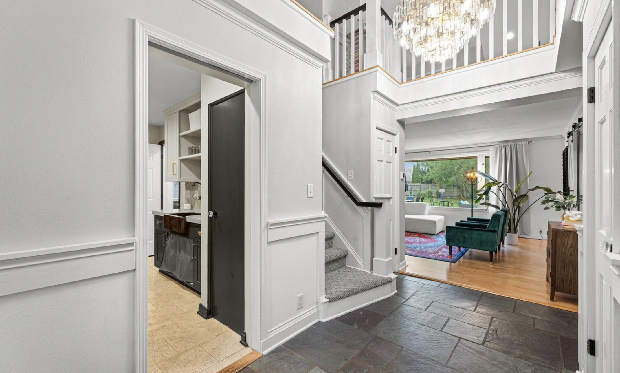 Interior view of a modern home with a staircase leading to an upper floor, a living room with a large window, a chandelier, and various furnishings and decor.