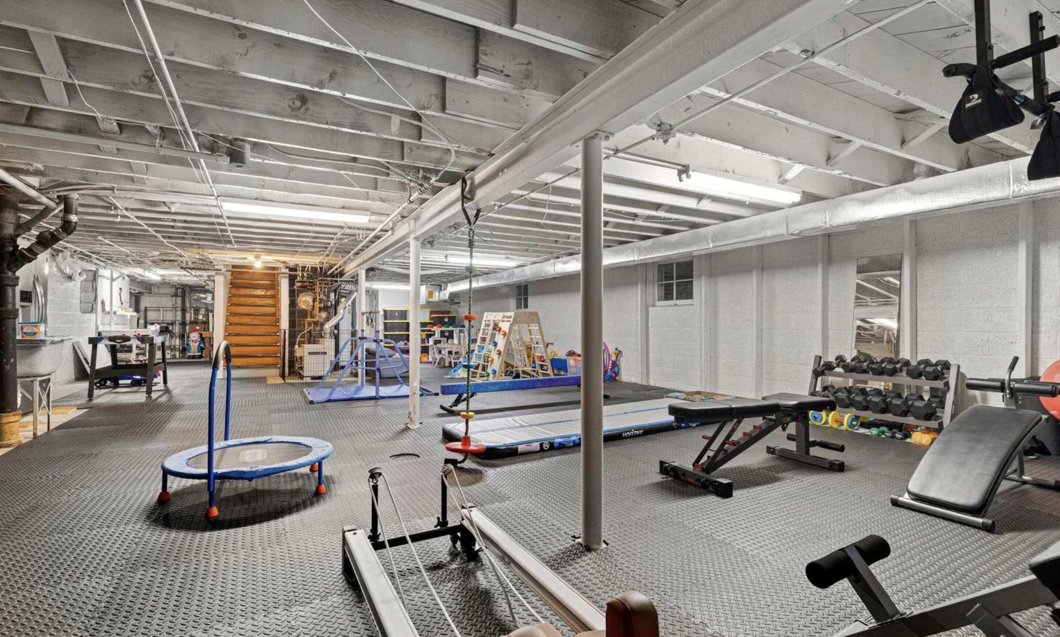 An indoor gymnasium and play area with fitness equipment, children's toys, and a trampoline, set in a basement with an unfinished ceiling and exposed pipes.