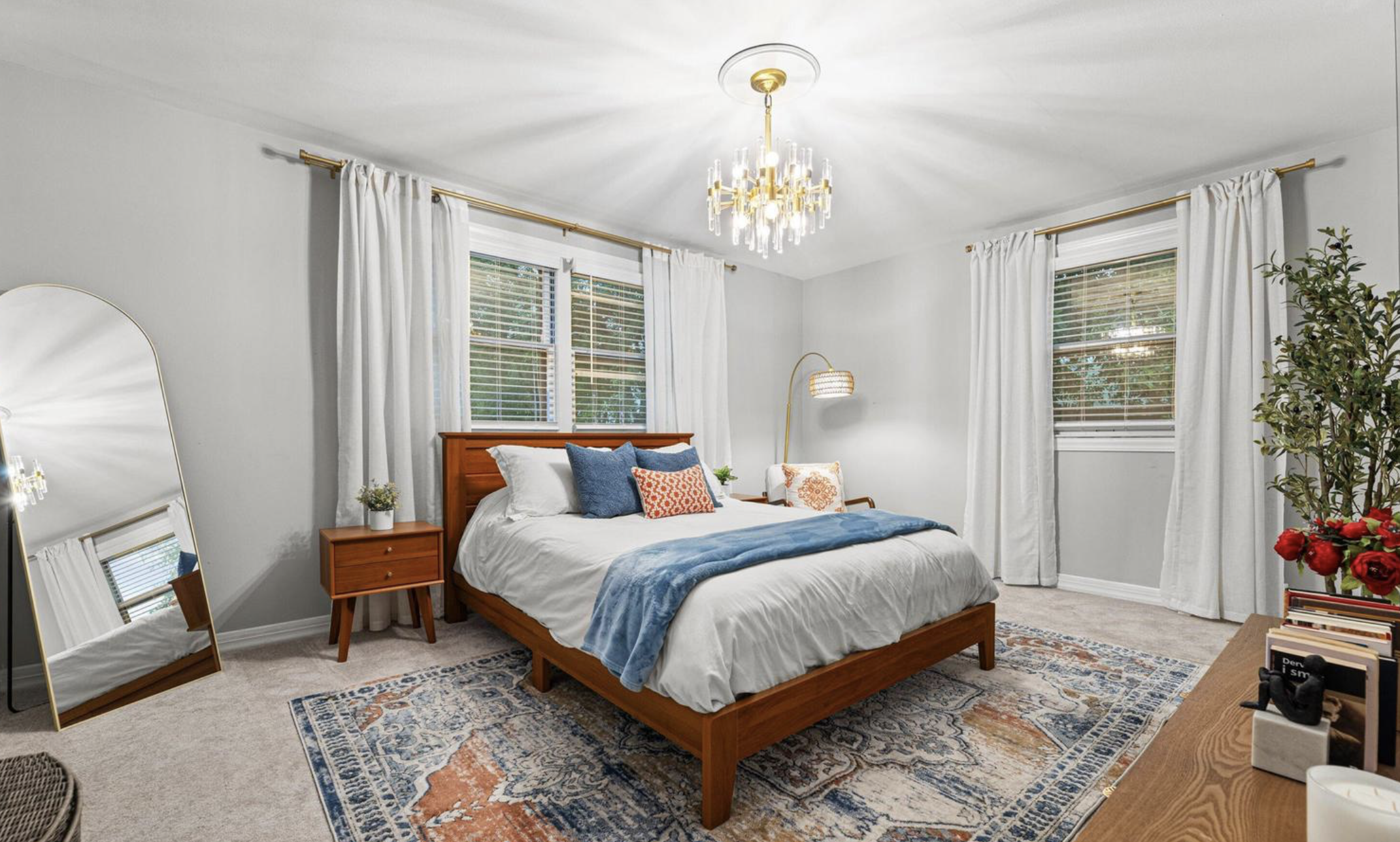 A bedroom with a wooden bed frame, white bedding, and blue and orange pillows. There are three windows with white curtains, a chandelier hanging from the ceiling, a floor lamp, a mirror, a nightstand, a colorful rug, and a plant in the corner.