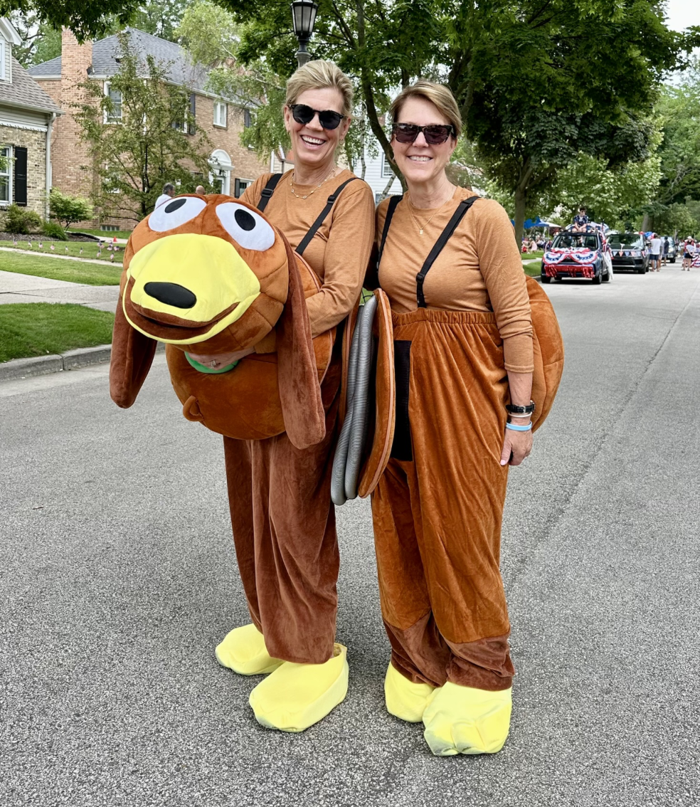 Two women dressed in matching brown outfits with yellow shoe coverings and sunglasses, standing on a neighborhood street during a parade. One woman is holding a large, plush mascot head resembling a cartoon dog with a big yellow nose and wide eyes.
