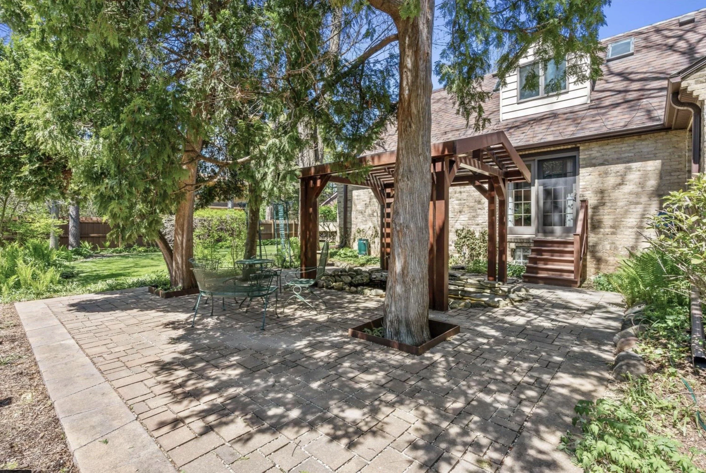 Backyard patio area with brick pavers, shaded by trees, with a wooden pergola, outdoor furniture, and a house with brick and siding in the background.