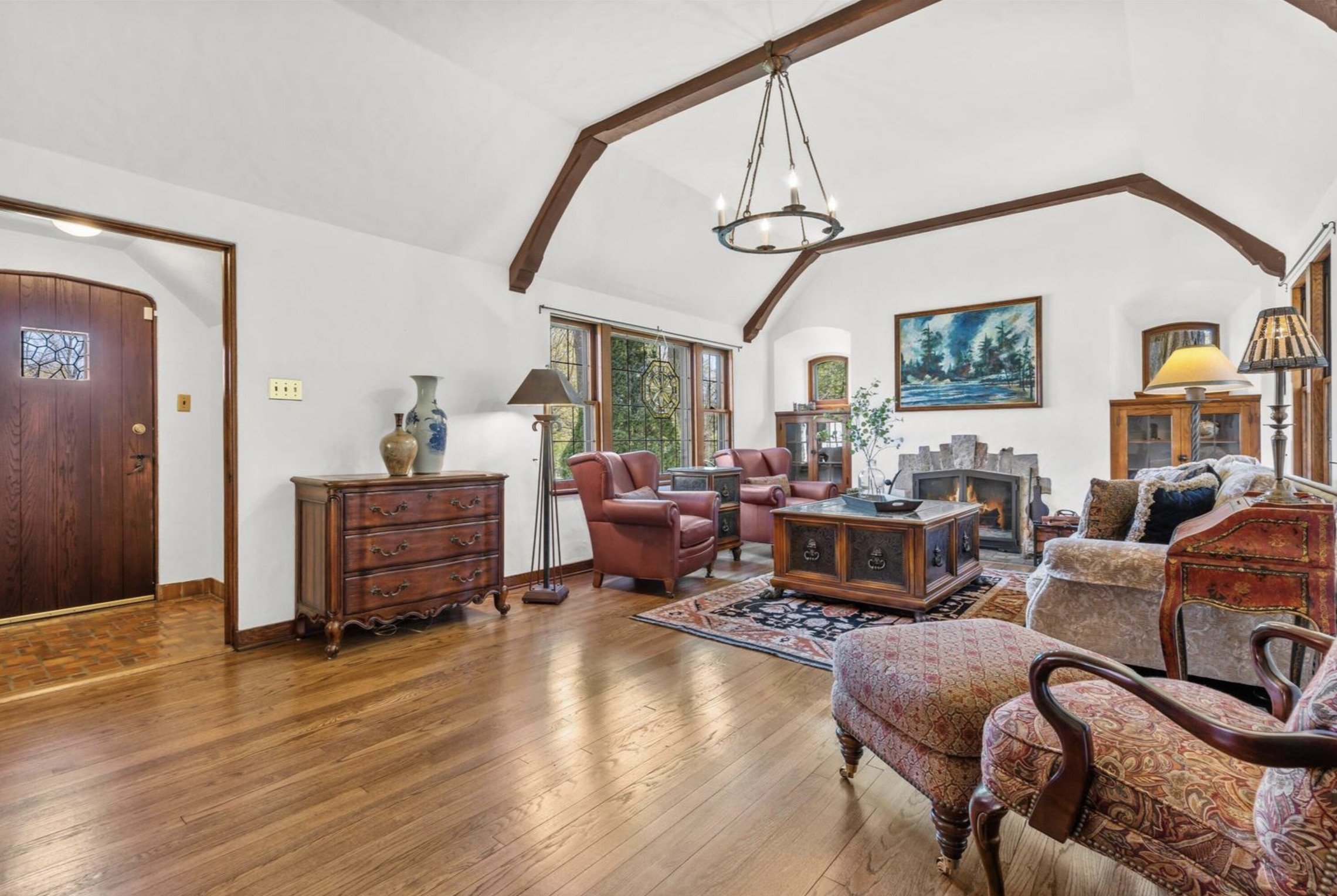 Living room with wooden floors, a fireplace, framed landscape painting, leather armchairs, a wooden coffee table, and large windows with greenery outside.
