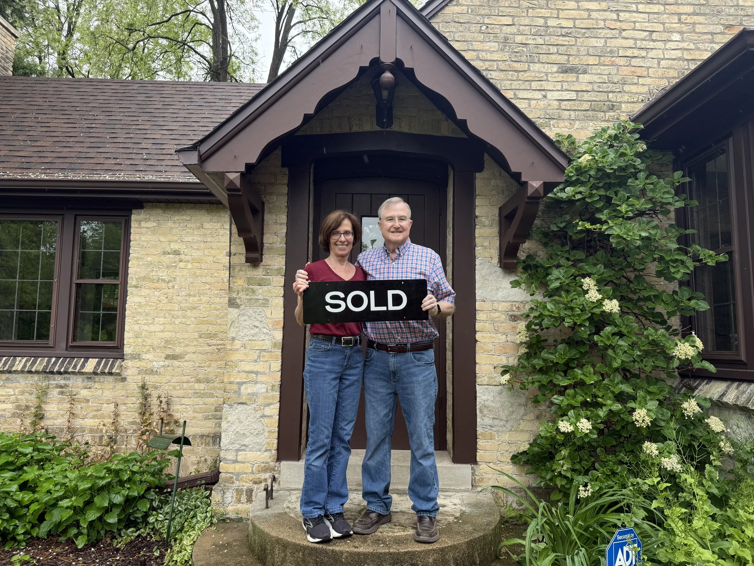 A smiling couple standing on a stone step in front of a house, holding a black "SOLD" sign, surrounded by green plants and flowers. The house has yellow brick walls and dark brown window frames with a decorative brown wooden canopy over the door.