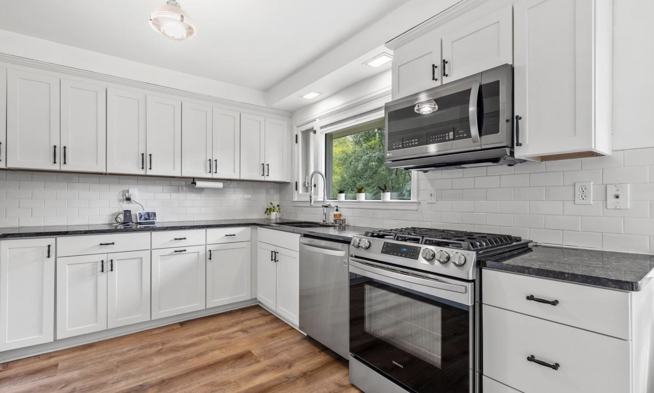 Kitchen with white cabinets, stainless steel microwave, oven, and dishwasher, white subway tile backsplash, black granite countertops, single-basin sink, window with greenery outside, hardwood floor.