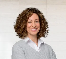Smiling woman with curly hair wearing a gray sweater and white shirt, standing in front of a light-colored wooden background.