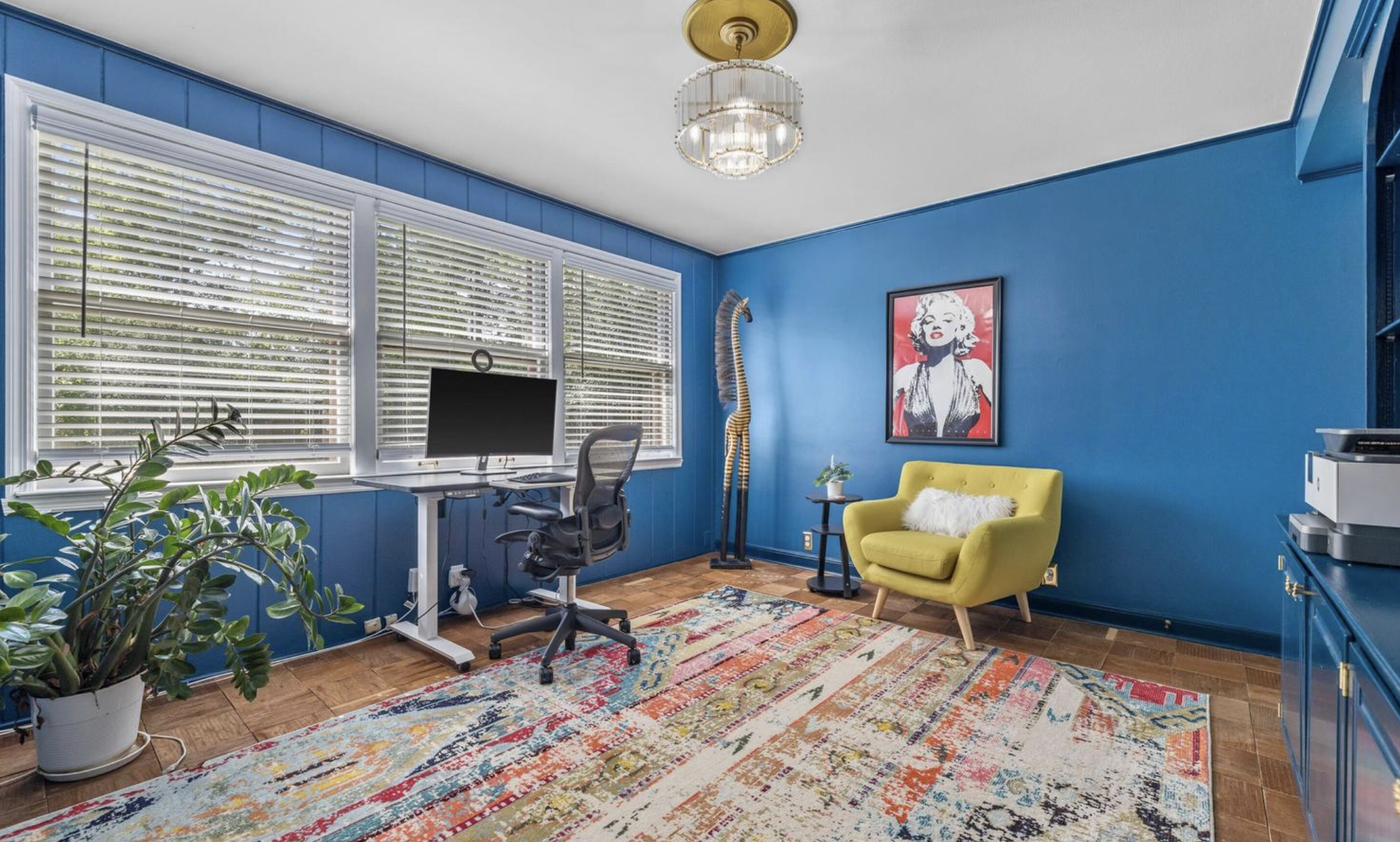 A brightly colored home office featuring blue walls, a large window with white blinds, a black office chair, a white desk with a computer monitor, and a yellow armchair with a white pillow. The room has a colorful patterned rug, framed Marilyn Monroe artwork, and decorative elements like a plant and a giraffe sculpture.