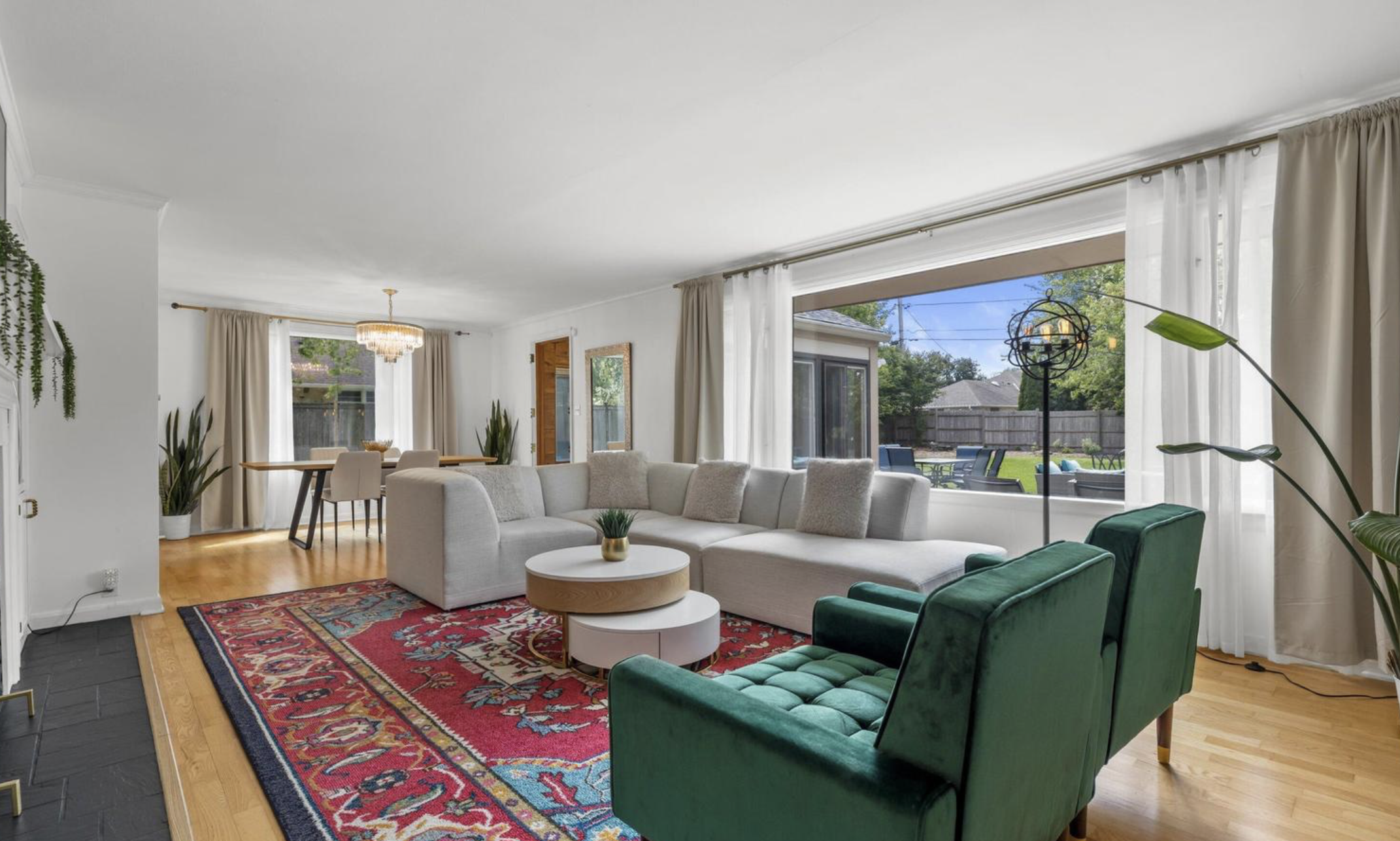Living room with large window, white sectional sofa, green armchairs, red patterned rug, wooden coffee table, dining area with chandelier, and outdoor patio visible through window.