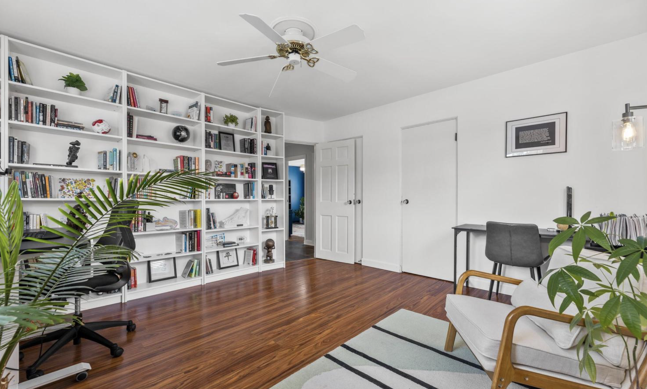 Living room with white bookshelves, a gray ceiling fan, wooden floor, white walls, framed artwork, a table with a black chair, a white armchair with wooden armrests, and several green plants.