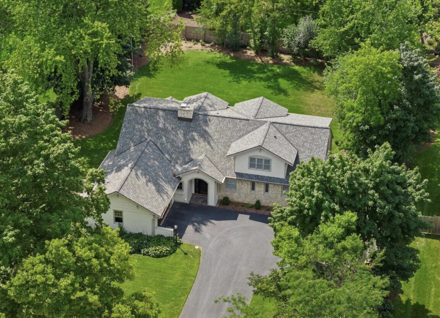 An aerial view of a large house with a grey shingle roof surrounded by tall green trees, a green lawn, and a curved driveway leading to the front entrance.